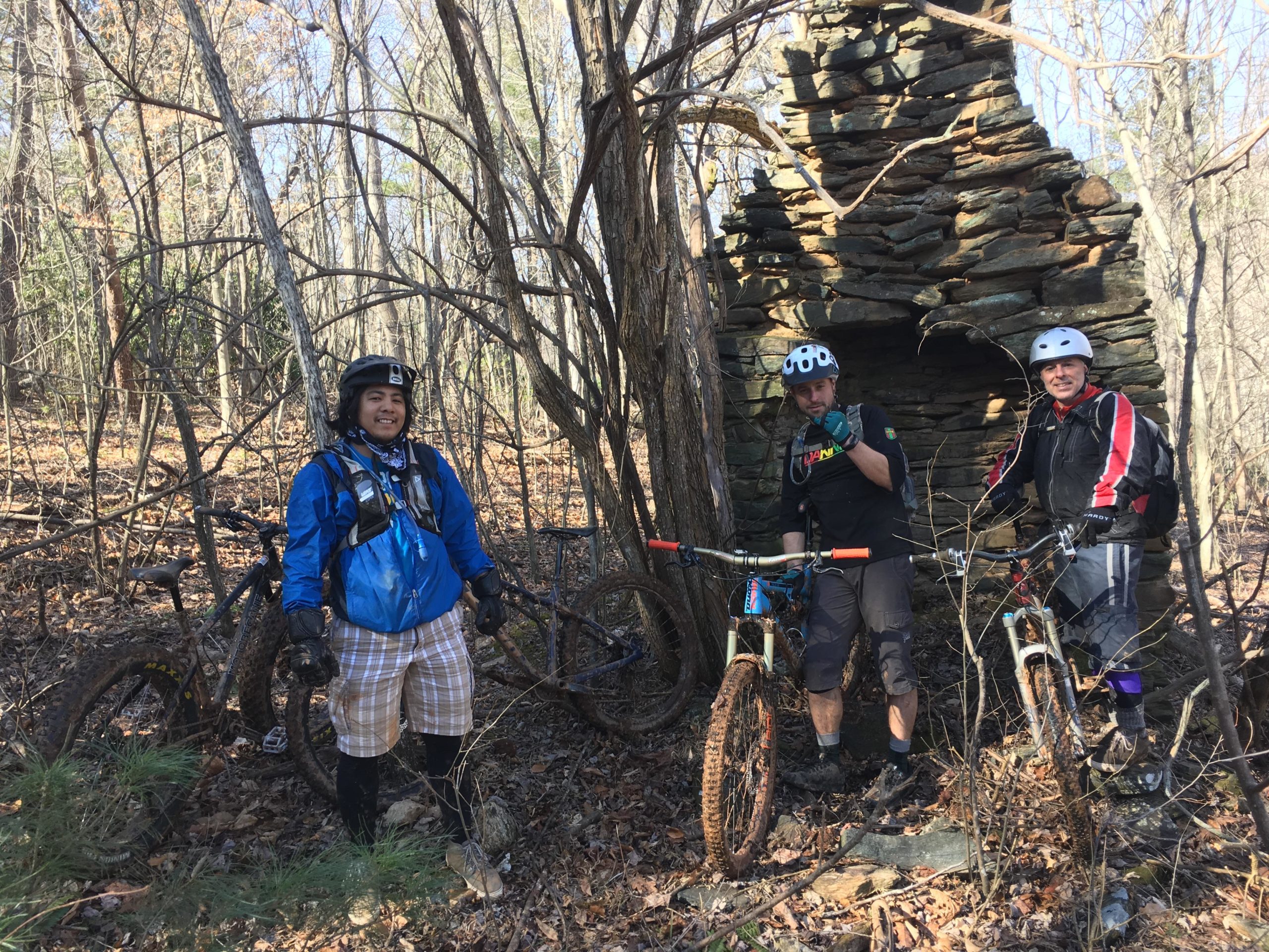 Three mountain bikers pose in a wooded area, standing next to an old stone structure partially covered by trees. The first person, wearing a blue jacket and plaid shorts, smiles at the camera. The second biker, in a green shirt and helmet, gives a thumbs-up. The third, dressed in a black and red outfit, stands next to their bike, all surrounded by fallen leaves and branches. Woolwine Trails [Shiners Revenge] mountain bike trail.