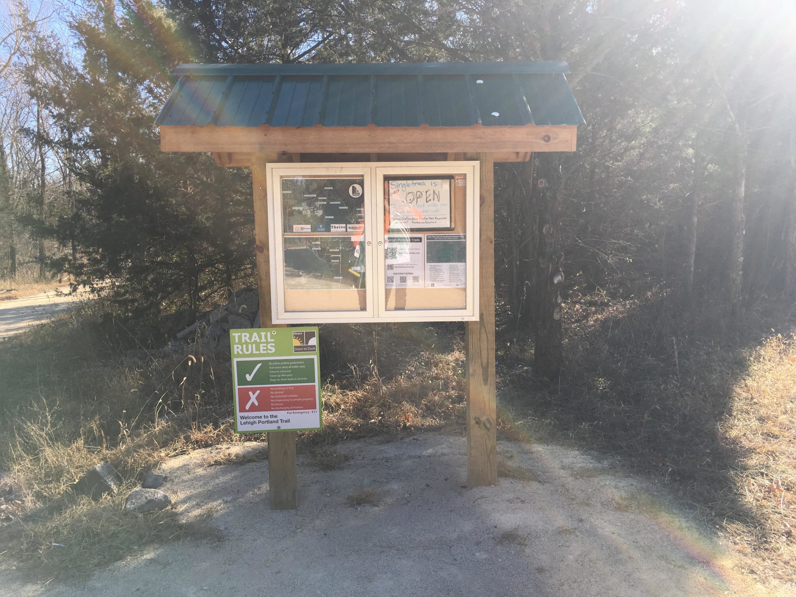 A wooden trail sign with a green roof displaying trail rules and maps. The sign has a notice that states "Trail is OPEN" and includes various informational flyers and guidelines. Surrounding the sign are trees and grassy areas, with a dirt path leading into the woods. Sunlight creates a lens flare effect in the upper right corner of the image. Lehigh Portland Trails mountain bike trail.