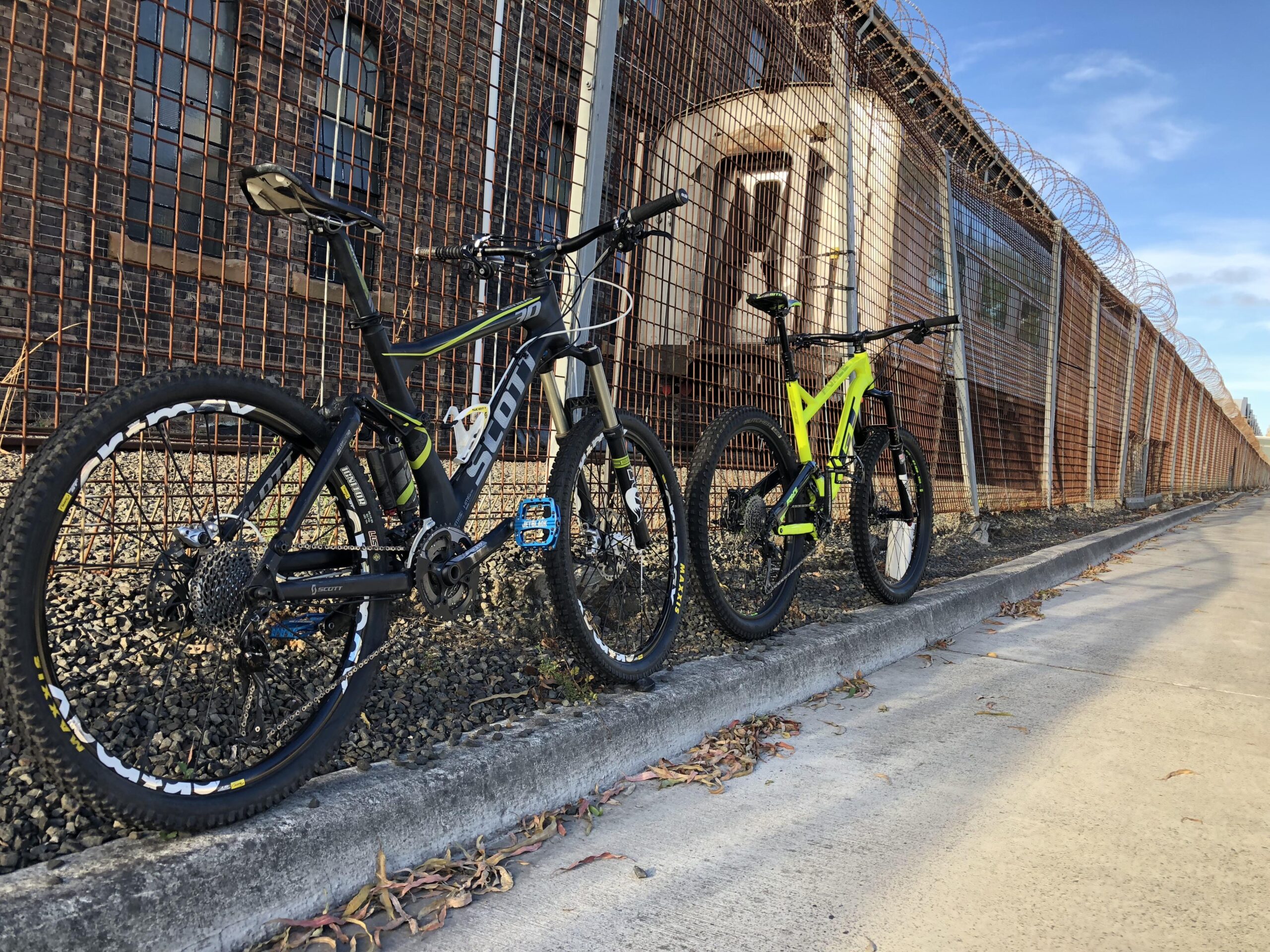 Scott Genius: Two mountain bikes are parked against a fence in an outdoor setting. The bike on the left has a black frame with green and blue accents, while the bike on the right is bright yellow. The background features a rusty metal fence with barbed wire and a building partially visible. The ground is made of gravel, and there are some fallen leaves scattered nearby. The sky is partly cloudy.