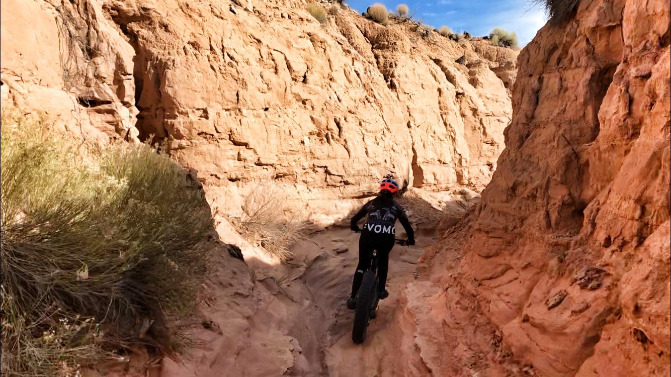A cyclist riding a fat bike along a narrow dirt trail surrounded by towering rock walls, with golden sagebrush visible on the side. The terrain is sandy and rugged, and the sky above is clear with a few clouds. The cyclist is wearing a helmet with a bright orange visor and a black jersey featuring the word "VOMO." Mariposa Fat Bike Trails mountain bike trail.