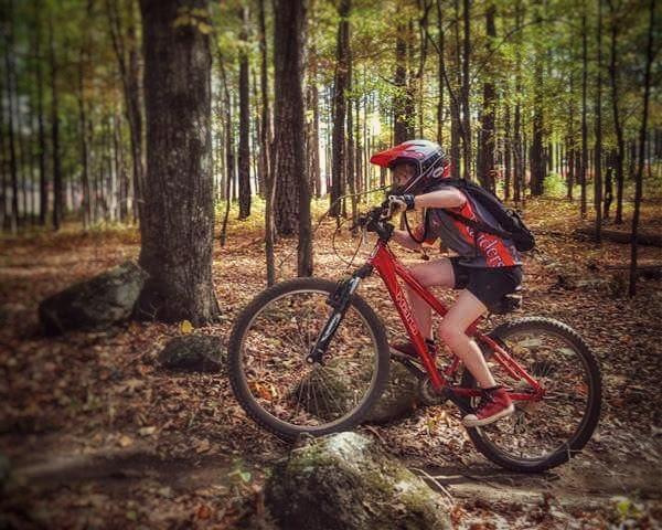 A young girl wearing a helmet rides a red mountain bike over rocky terrain in a wooded area. The autumn foliage surrounds her, creating a vibrant landscape of orange and yellow leaves. Dauset Trails Nature Center mountain bike trail.
