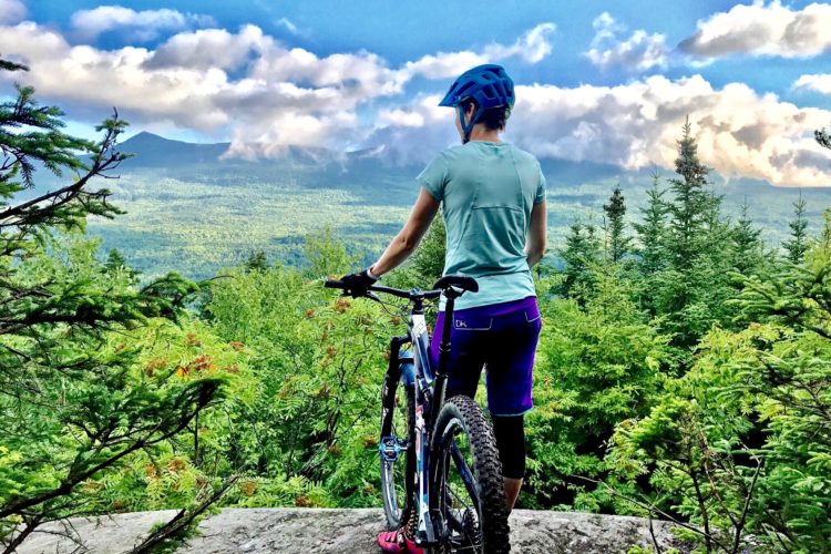 A person standing on a rock ledge, facing away and looking out at a lush green landscape with mountains in the distance. They are wearing a blue helmet and a light blue shirt, with purple shorts and gloves. A mountain bike stands beside them, surrounded by trees and under a partly cloudy sky.