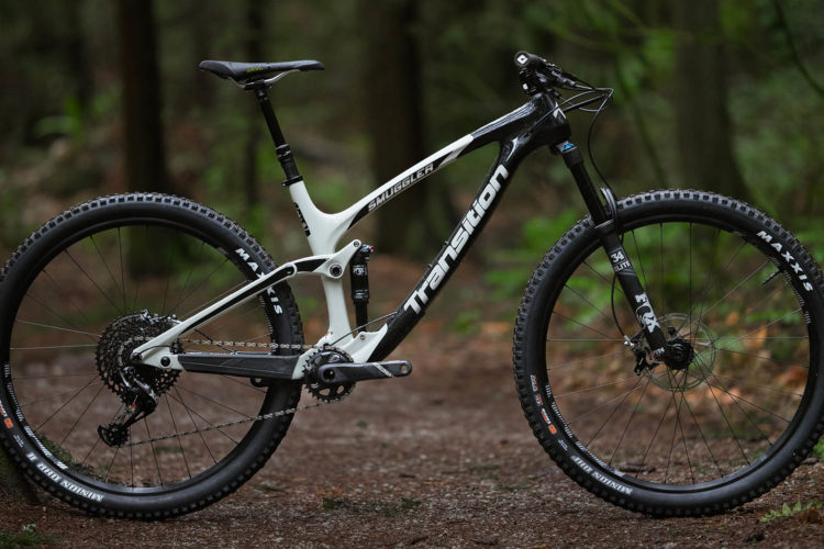 A black and white mountain bike is positioned on a forest trail, surrounded by trees and greenery. The bike features thick tires, a lightweight frame, and advanced suspension components, showcasing its design for rugged terrain and off-road cycling.