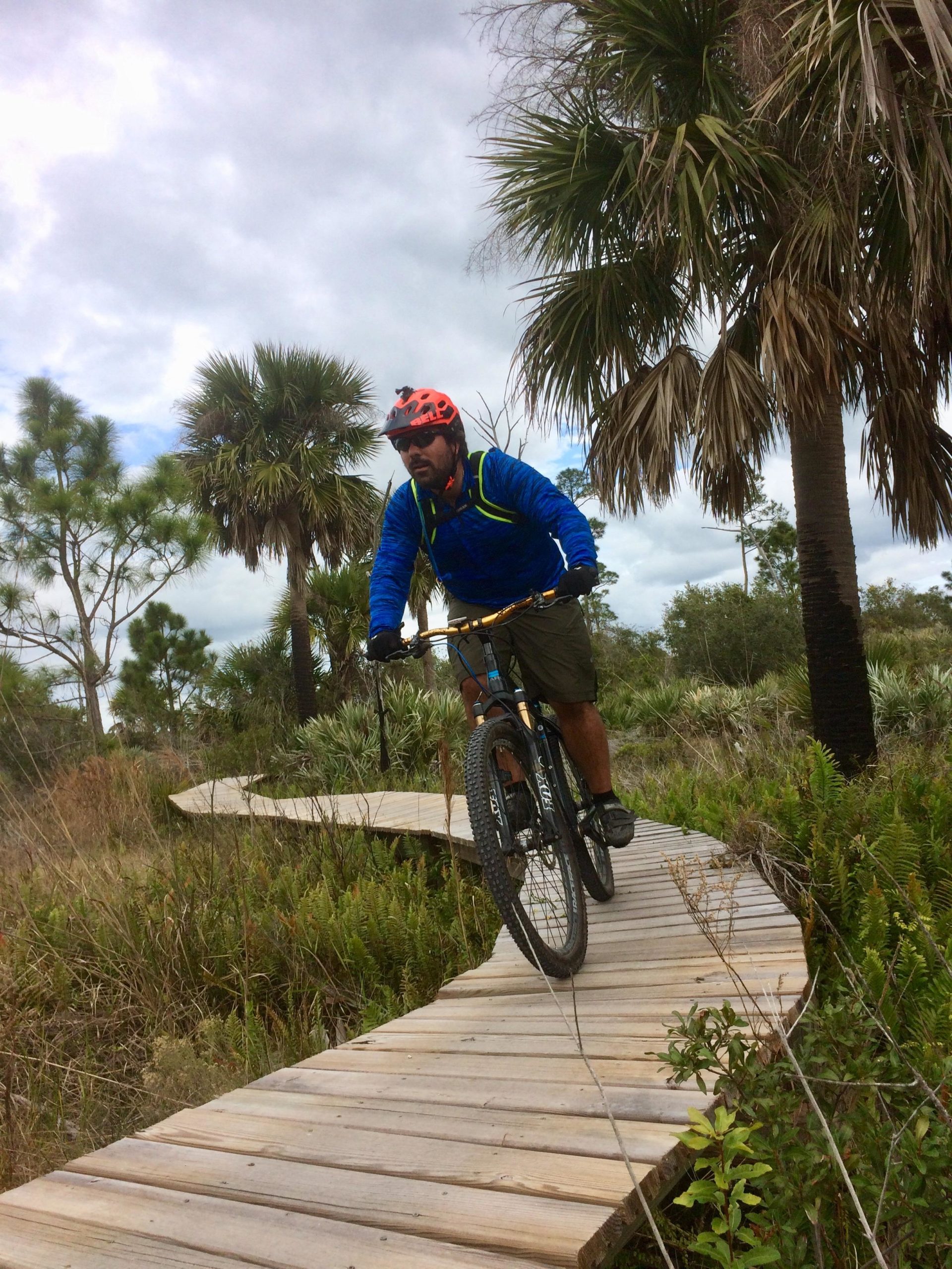 A person wearing a blue jacket, helmet, and shorts rides a mountain bike on a wooden boardwalk surrounded by greenery and palm trees under a cloudy sky. Jonathan Dickinson State Park mountain bike trail.