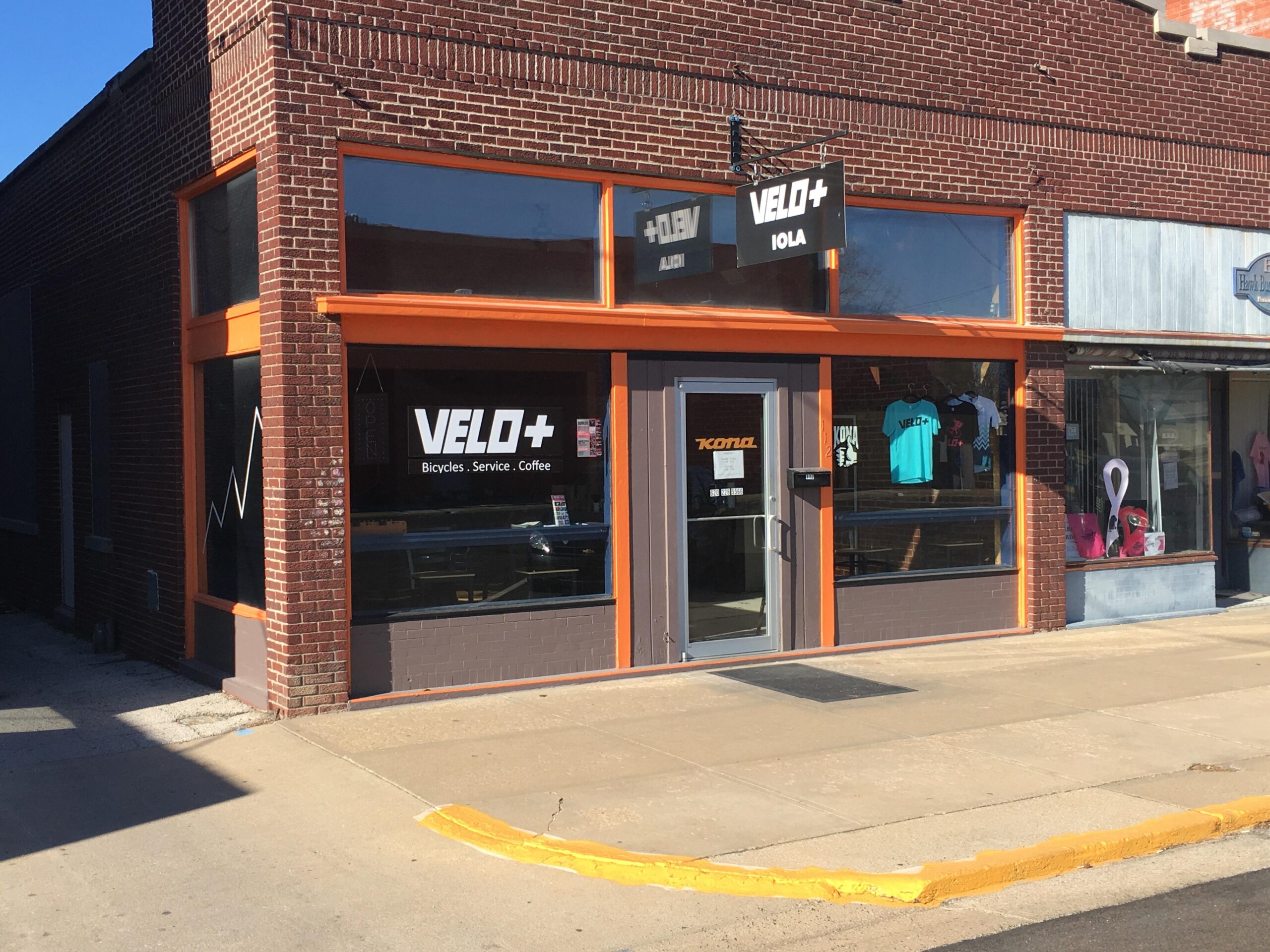 Image of a brick storefront for Velo+, a shop that offers bicycles, service, and coffee. The front features large windows with orange trim, displaying bicycles and merchandise inside. A sign above the entrance reads "VELO+ IOLA," and an "OPEN" sign is visible in the window. The building is situated on a city street with a yellow curb in front.