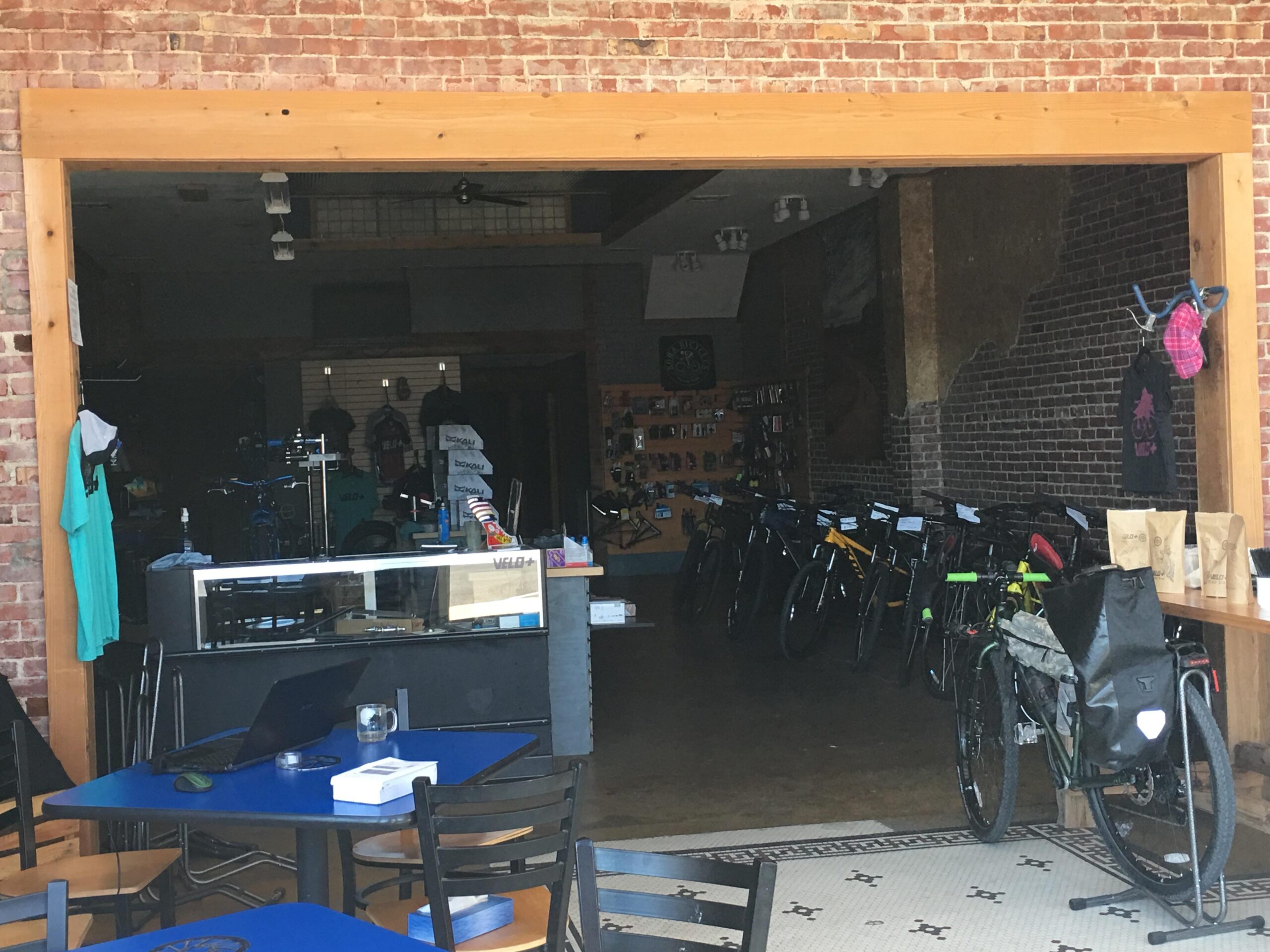 Alt text: View of a bicycle shop from an outdoor seating area, featuring a display of bicycles and cycling gear inside. A blue table with a laptop is set up in the foreground, with several bicycles lined up along the back wall. The shop has a rustic brick interior and wooden accents.