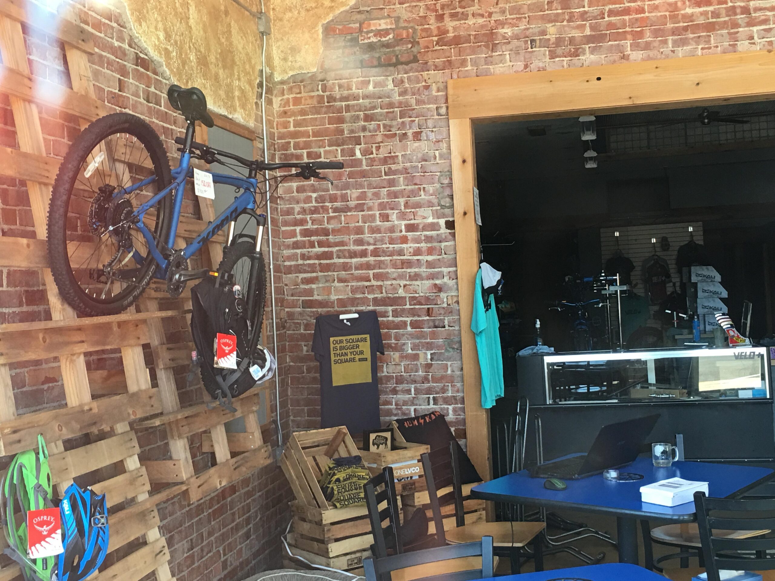 A blue mountain bike is mounted on a wooden display against a brick wall inside a shop. Below the bike, a black backpack with tags hangs. There are several helmets in green and blue colors on the display, and a table with a laptop and office supplies is visible in the foreground. In the background, various biking apparel can be seen hanging, along with another bicycle and some crates.