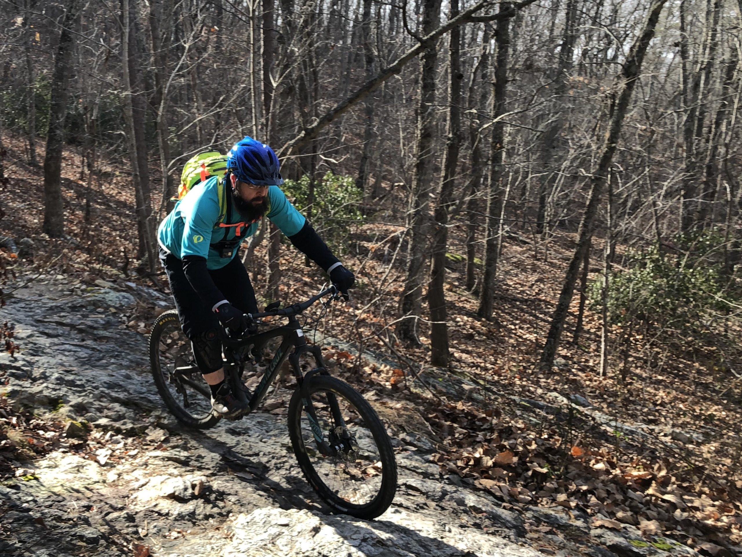 A mountain biker navigates a rocky trail in a wooded area during the daytime, wearing a blue helmet and a bright turquoise shirt, with a backpack on. The ground is covered in leaves and rocks, surrounded by bare trees, indicating a winter setting. Uwharrie NF: Wood Run, Supertree And Keyauwee mountain bike trail.