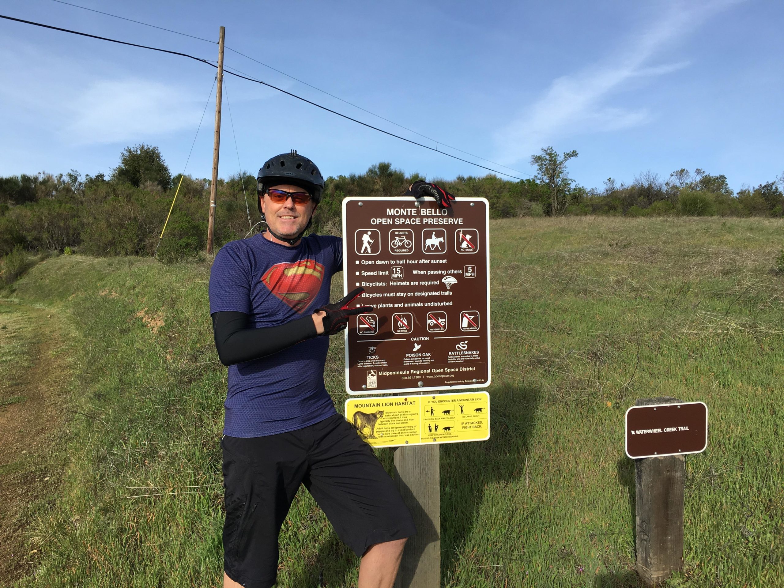 A person wearing a helmet and sunglasses stands next to a sign at Monte Bello Open Space Preserve, which provides information about park rules and safety guidelines. The background features green hills and blue skies, indicating a sunny day. Monte Bello / Rancho San Antonio mountain bike trail.