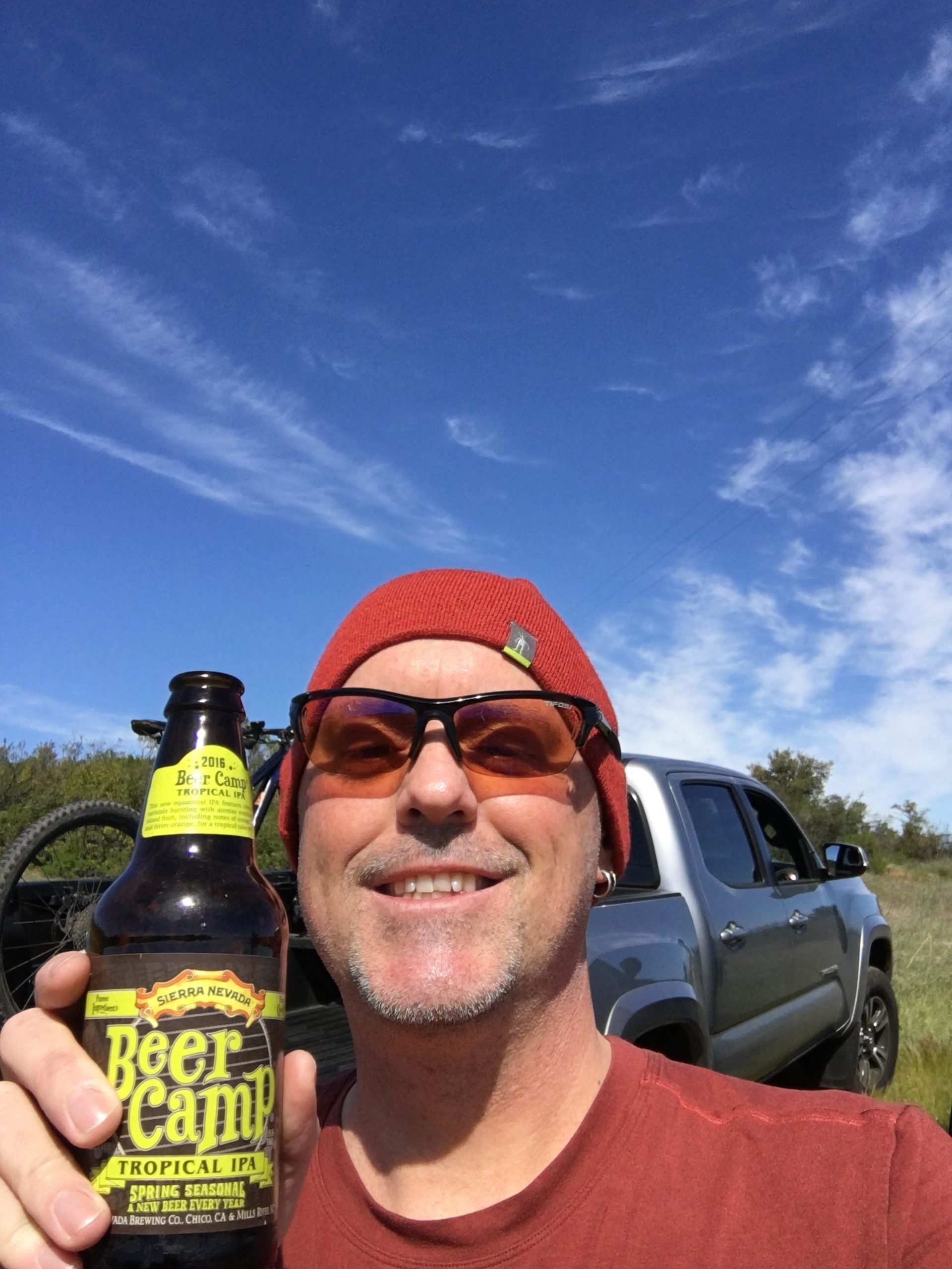 A person wearing sunglasses and a red beanie smiles while holding a bottle of Beer Camp Tropical IPA. In the background, there is a gray pickup truck and a clear blue sky with wispy clouds. The setting appears to be outdoors, possibly in a rural area. Monte Bello / Rancho San Antonio mountain bike trail.