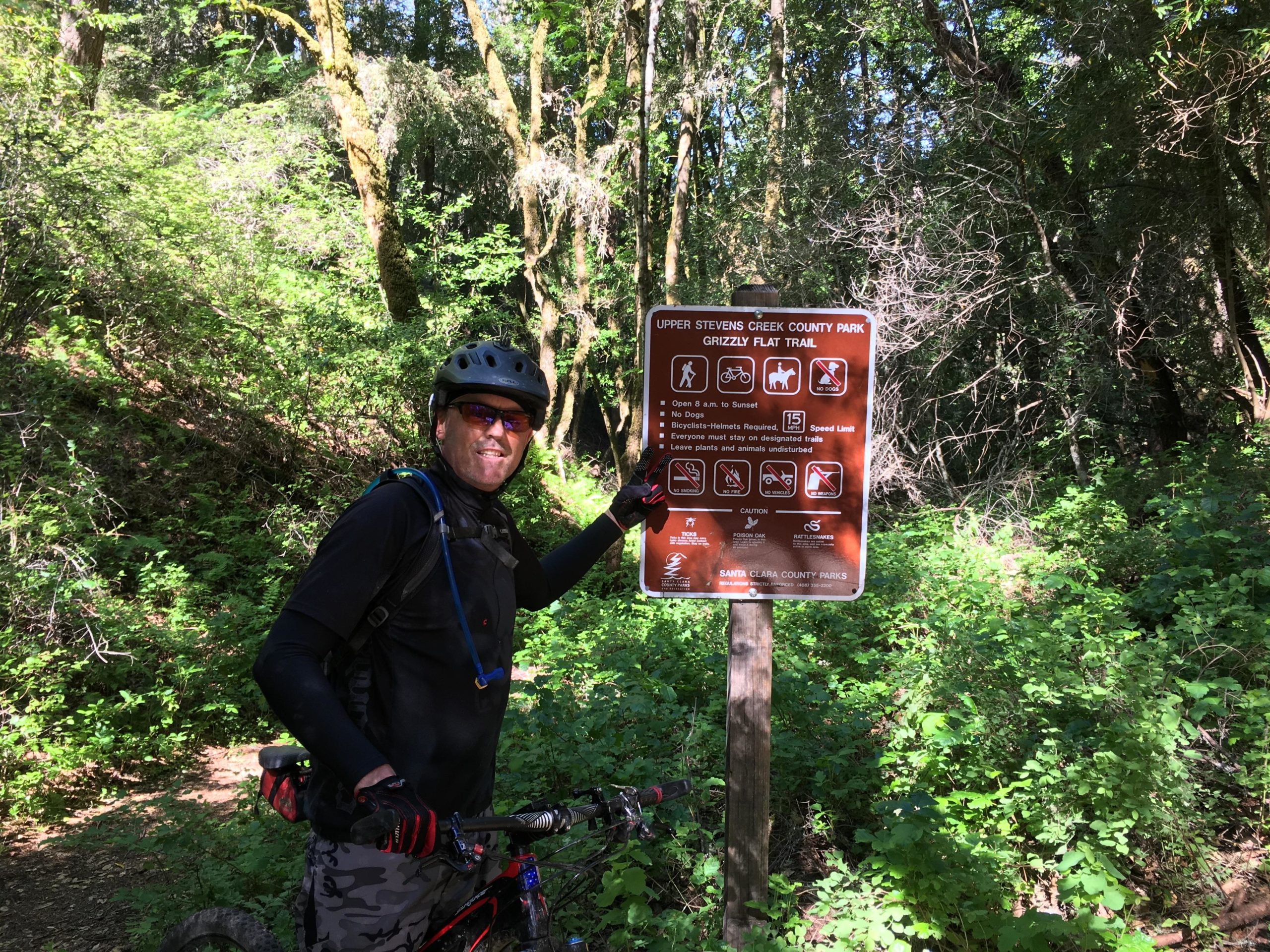 A cyclist in a black outfit and helmet stands next to a trail sign at Upper Stevens Creek County Park, Grizzly Flat Trail. The sign displays park regulations, including hours of operation, safety guidelines, and information about trail usage. Surrounding vegetation is lush and green, indicating a natural forested area. Saratoga Gap mountain bike trail.