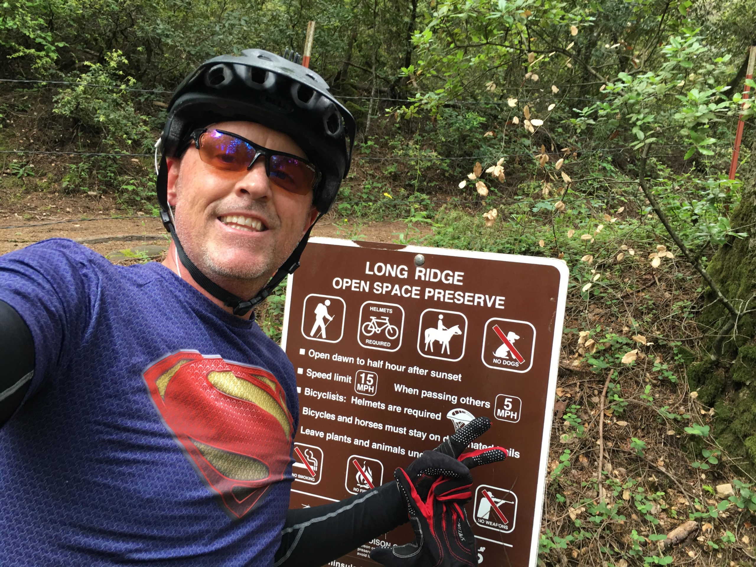A cyclist in sunglasses and a superhero-themed shirt smiles at the camera while standing near a trail sign in a wooded area. The sign provides information about the Long Ridge Open Space Preserve, including rules for bicyclists, speed limits, and guidelines for trail use. Saratoga Gap mountain bike trail.