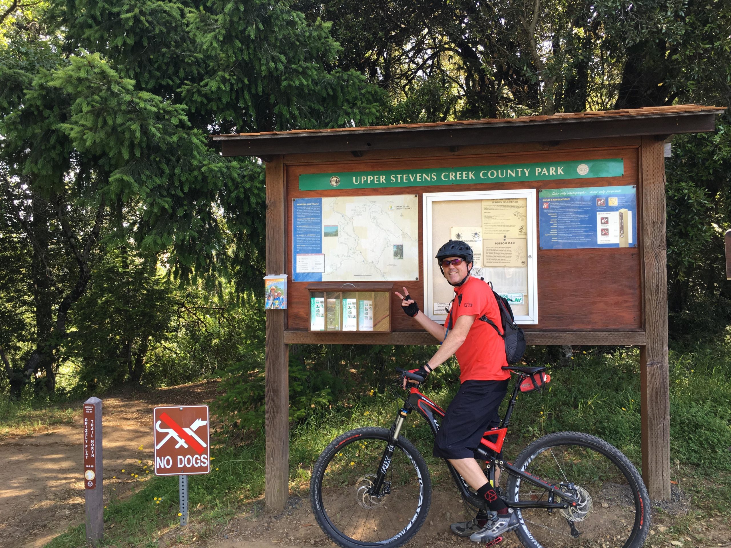 A cyclist in a red shirt and black shorts is standing next to a park information board at Upper Stevens Creek County Park. The board displays a map and various park guidelines. A "No Dogs" sign is visible nearby, along with a path leading into the park, surrounded by greenery. Saratoga Gap mountain bike trail.