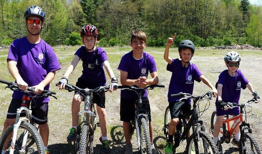 Trek Excalibur: A group of five young cyclists wearing purple t-shirts and helmets pose with their bikes in a grassy area surrounded by trees. The scene is bright and sunny, conveying a cheerful and active atmosphere.