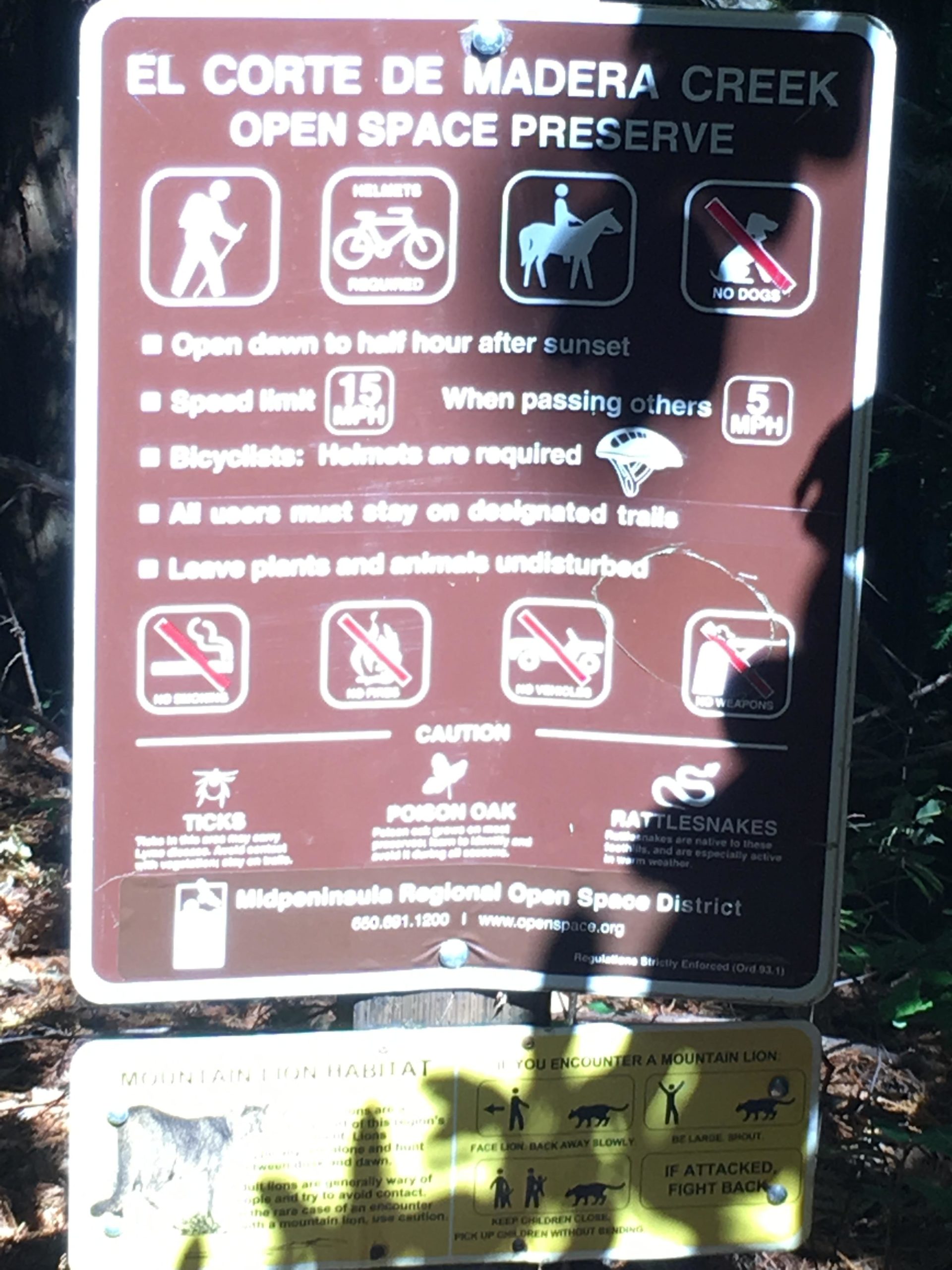 A trail sign for the El Corte de Madera Creek Open Space Preserve, featuring regulations and safety information for visitors. The sign includes icons for hiking, cycling, and horseback riding, along with rules such as helmet requirements, speed limits, and guidelines for staying on designated trails. It also contains warnings about wildlife, including mountain lions, ticks, and poison oak, plus advice for encounters with these animals. El Corte De Madera Creek Open Space mountain bike trail.