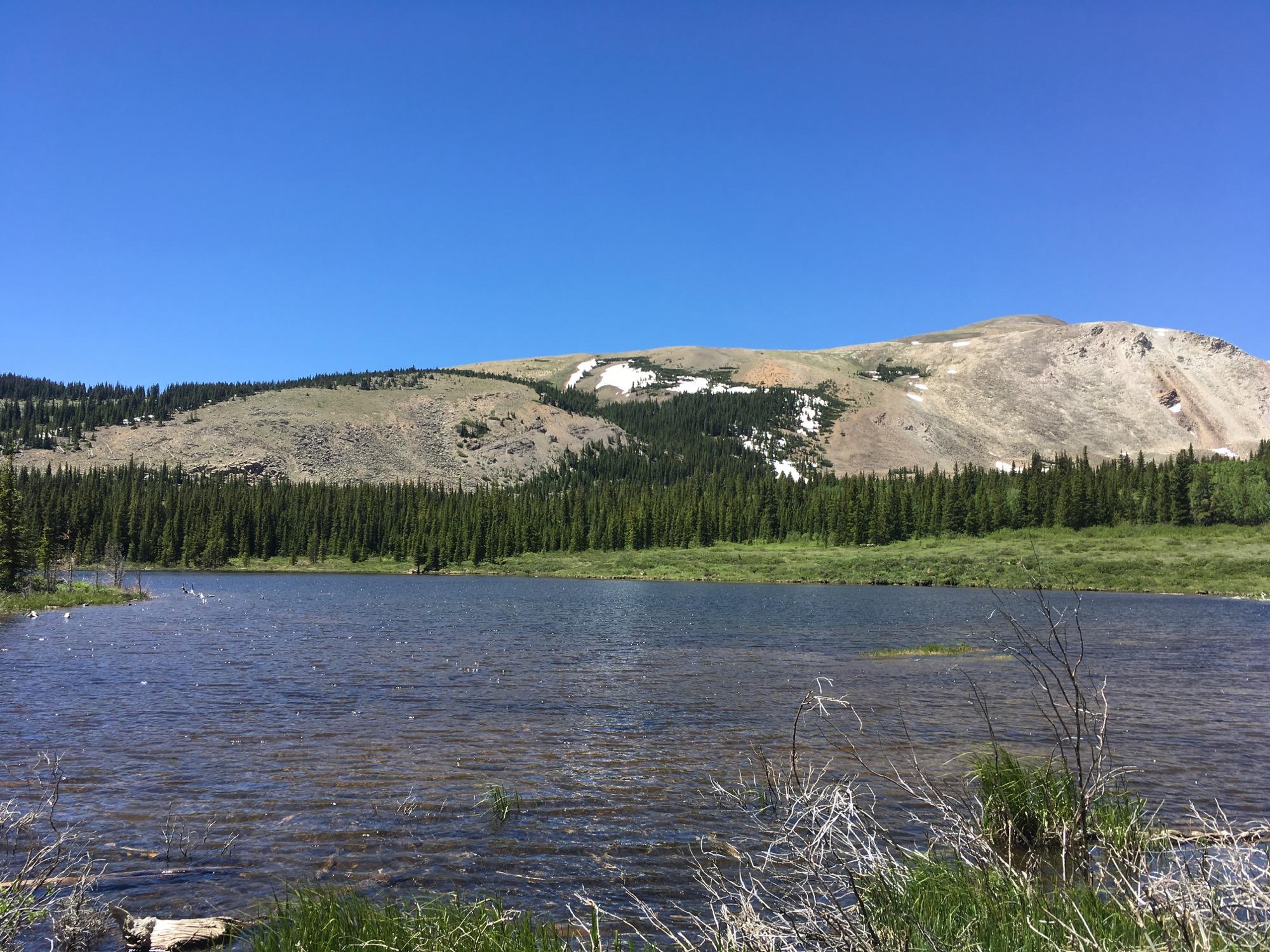 A serene landscape featuring a clear lake in the foreground, surrounded by lush green trees. In the background, a mountain with rocky slopes and patches of snow extends under a bright blue sky. The scene captures the tranquility of nature, with gentle waves on the water and greenery along the shore. Twelvemile Trail mountain bike trail.