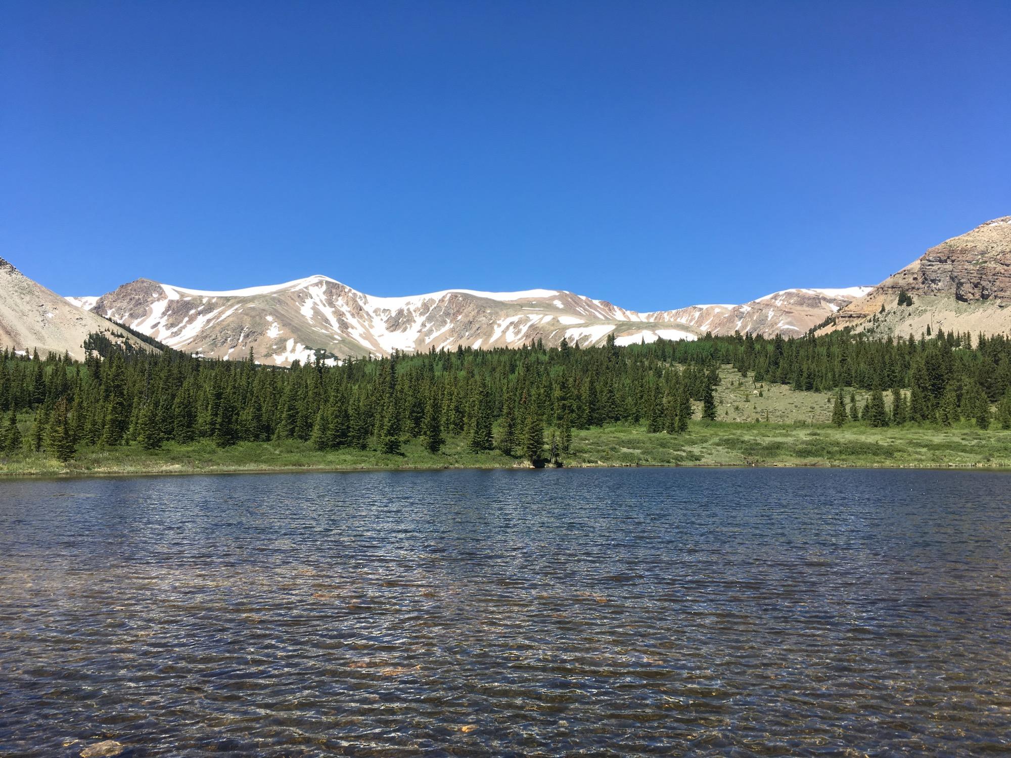 A serene landscape featuring a clear blue sky above snow-capped mountains. In the foreground, a calm lake reflects the surrounding greenery, with dense forests of trees lining the shore. The mountains in the background showcase varied textures and colors, creating a picturesque natural scene. Twelvemile Trail mountain bike trail.