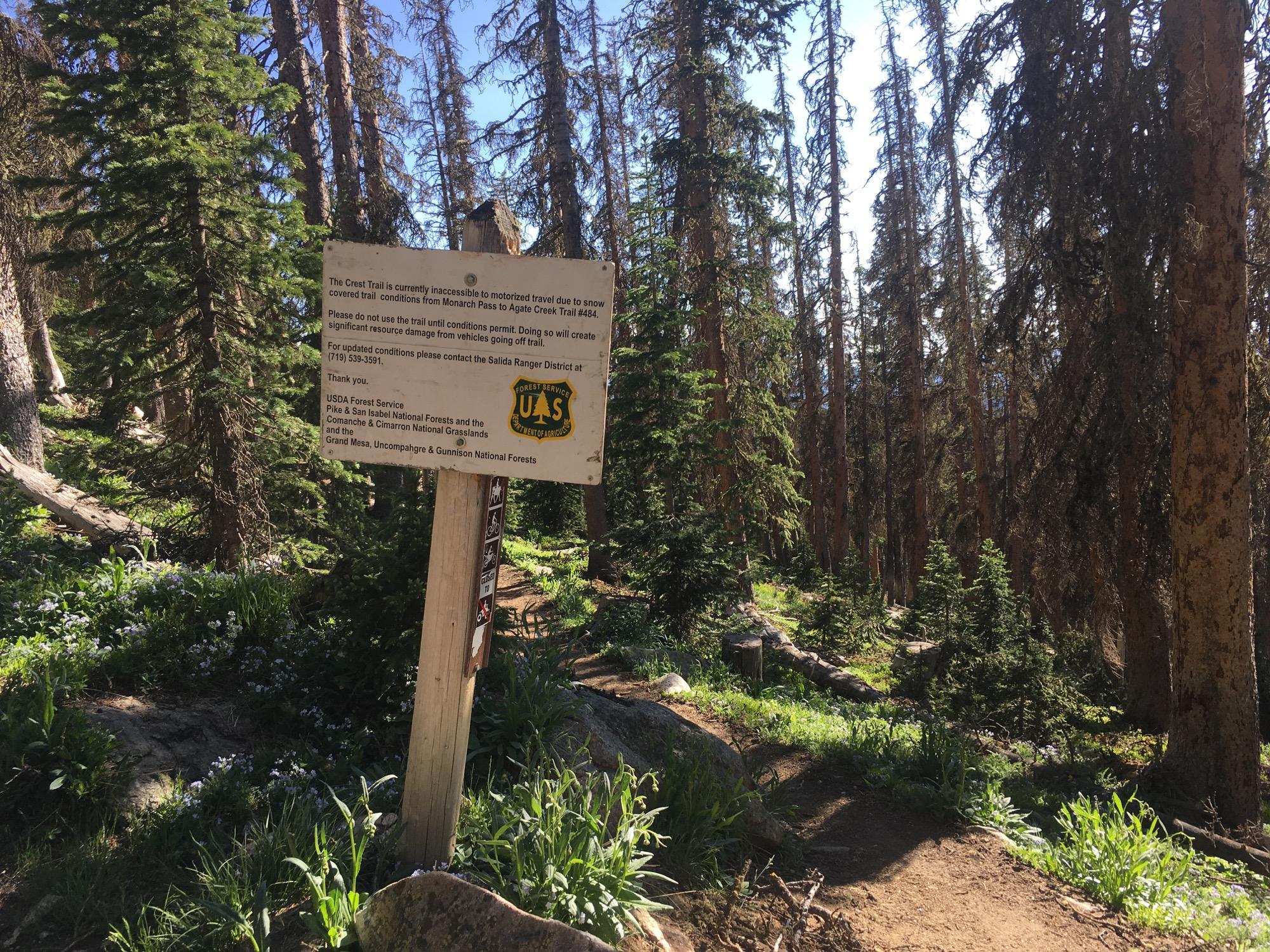 Sign in a forested area providing trail information. The sign indicates that the Crest Trail is currently inaccessible to motorized vehicles due to snow, advises against using the trail until conditions improve, and offers a contact number for updates. Surrounding the sign are tall trees and green vegetation, characteristic of a natural forest environment. Monarch Crest Trail mountain bike trail.