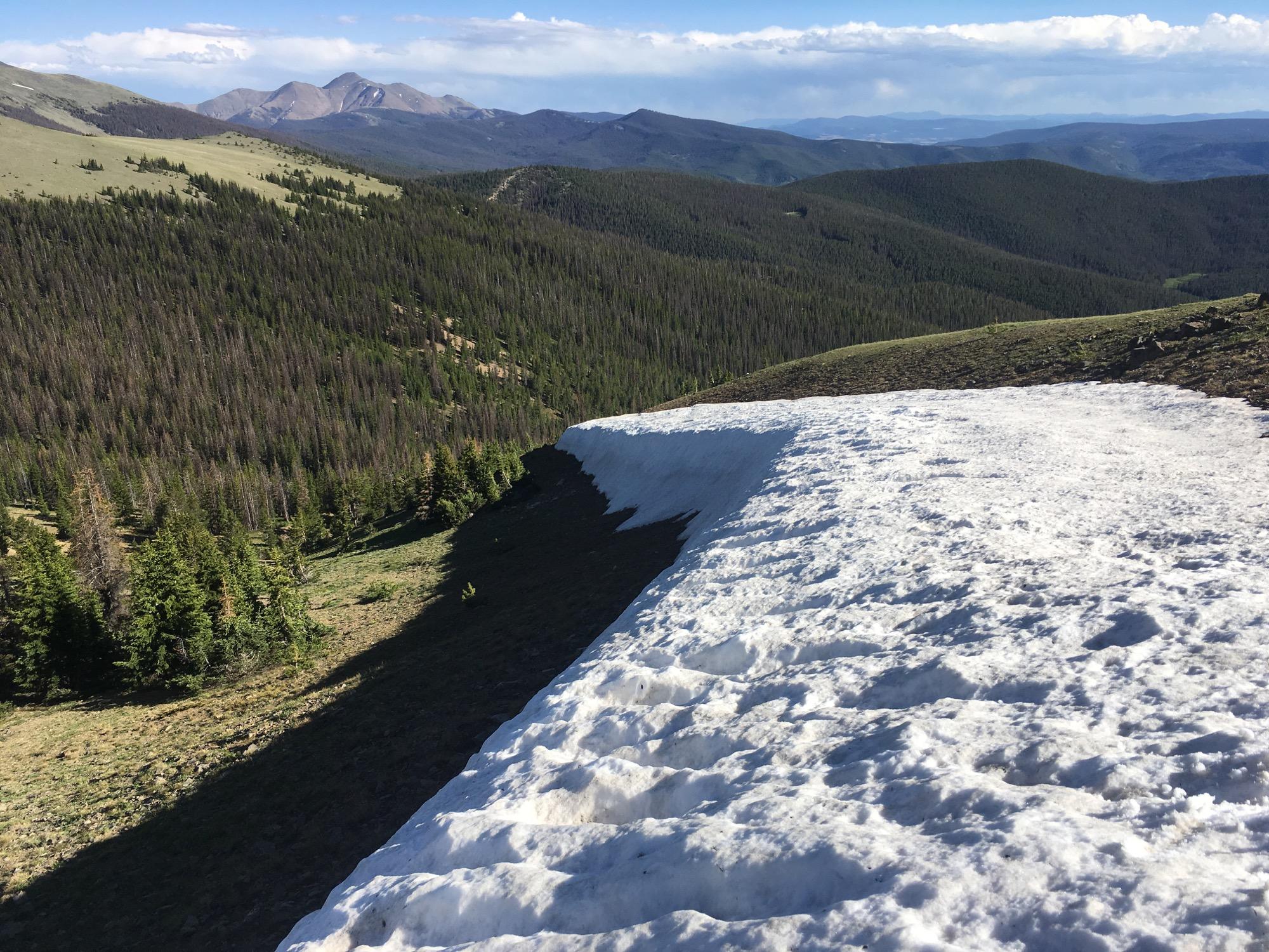 A panoramic view of a mountainous landscape featuring a snowy ridge in the foreground, with lush green forests below and distant peaks under a blue sky dotted with clouds. The scene captures the contrast between the snow and the greenery, highlighting the natural beauty of the area. Monarch Crest Trail mountain bike trail.