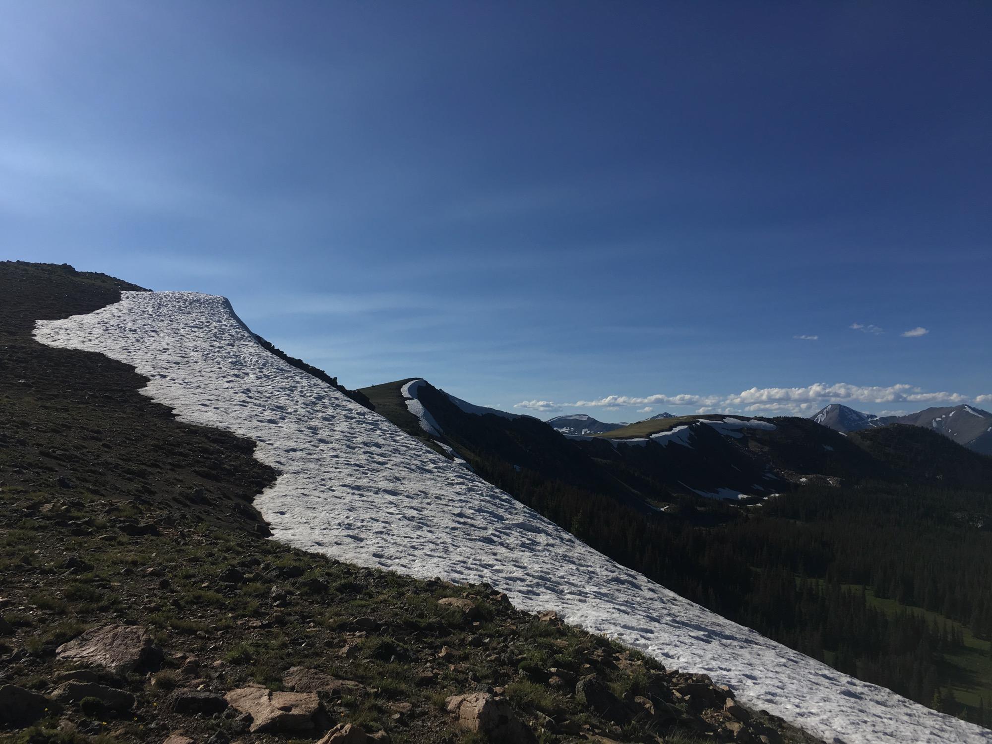 A panoramic view of a mountainous landscape featuring a snowfield that flows down the slope, surrounded by rocky terrain and lush greenery. The sky is clear with a few clouds, suggesting a bright day. In the background, additional mountains can be seen, some with patches of snow. Monarch Crest Trail mountain bike trail.