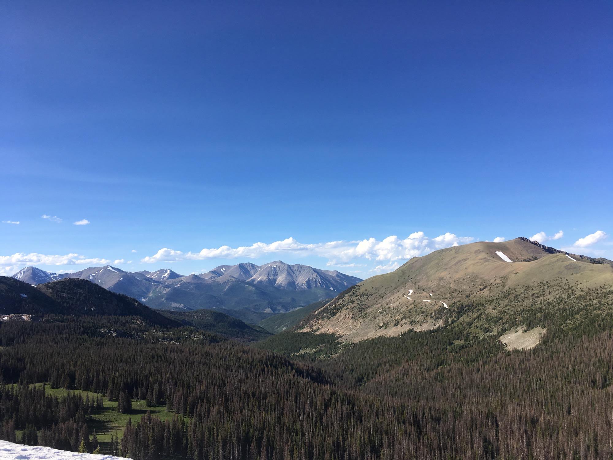 A scenic view of a mountainous landscape under a clear blue sky, featuring green valleys and patches of snow on the mountain peaks in the distance. The foreground showcases a dense forest of evergreen trees, while various mountain ranges rise majestically in the background. Monarch Crest Trail mountain bike trail.