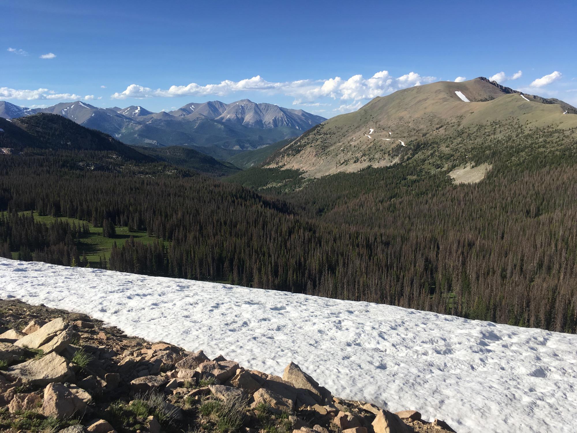 A panoramic view of a mountainous landscape featuring a snow-covered foreground, dense evergreen forests, and rolling hills in the background under a clear blue sky with scattered clouds. Monarch Crest Trail mountain bike trail.