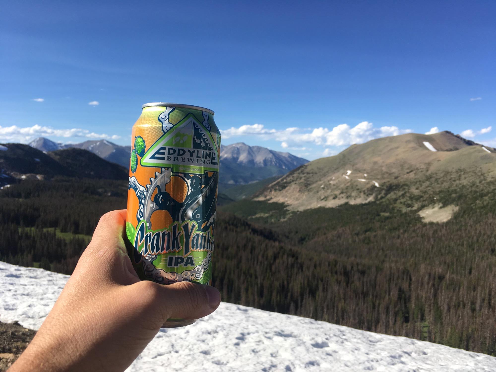 A hand holding a can of Eddyline Brewing's Crank Yanker IPA in front of a picturesque mountain landscape, featuring blue skies and distant peaks. Snow is visible in the foreground, indicating a high-altitude setting. Monarch Crest Trail mountain bike trail.