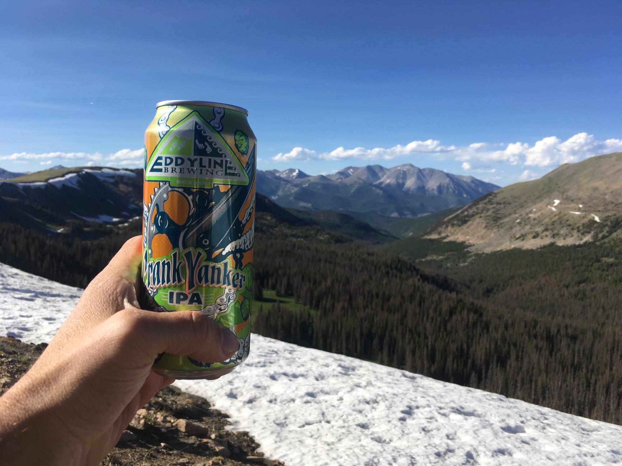 A hand holding a colorful can of Eddyline Brewing's Crank Yanker IPA, set against a scenic mountain backdrop featuring snow-capped peaks and lush green valleys under a clear blue sky. Monarch Crest Trail mountain bike trail.