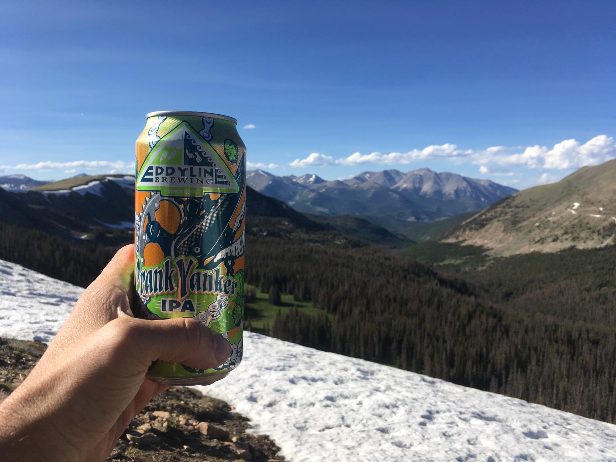 A hand holding a can of Eddyline Brewing's "Frank Yanker IPA" against a scenic mountain backdrop featuring snow-capped peaks and a clear blue sky. Monarch Crest Trail mountain bike trail.
