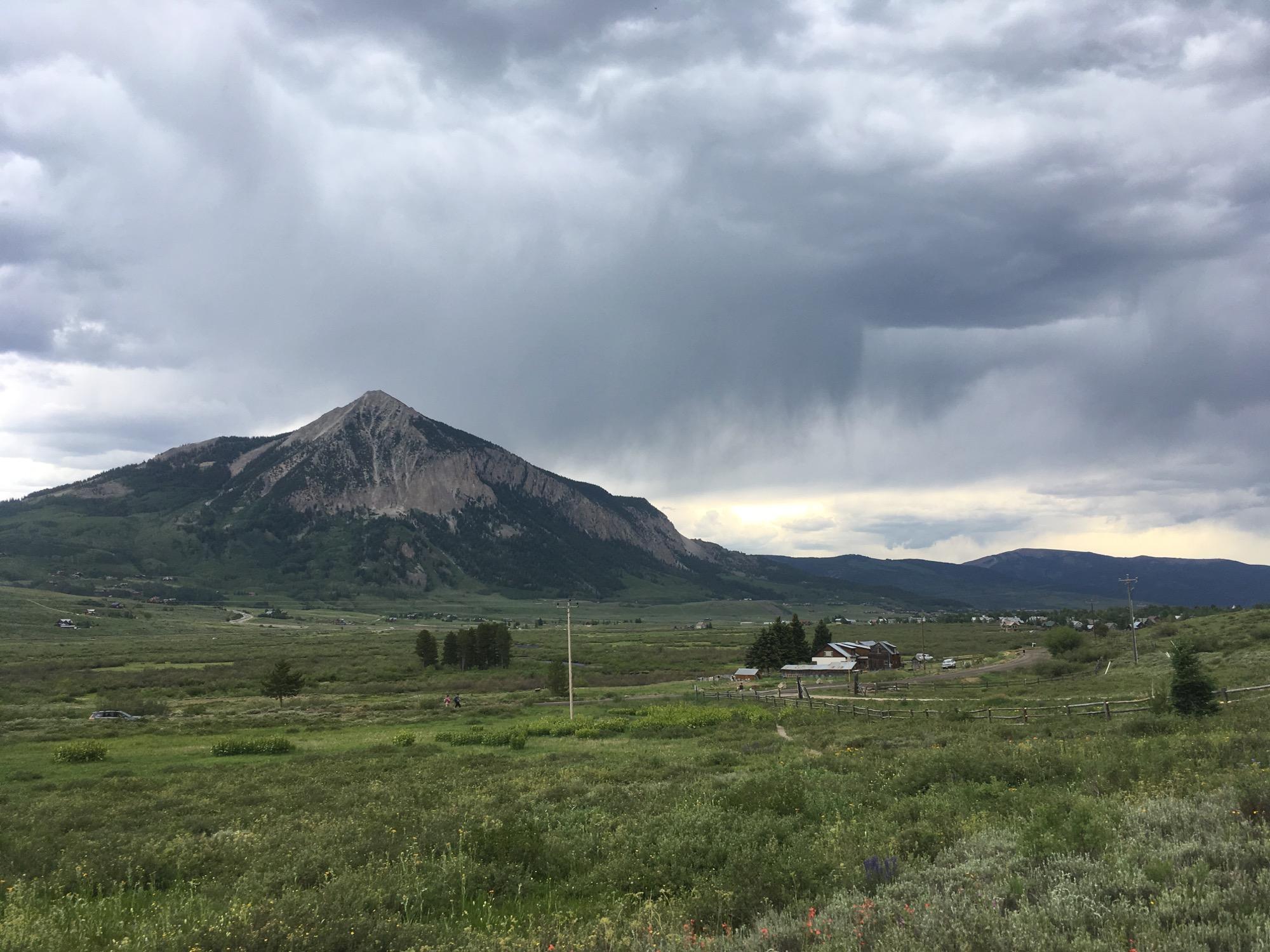 A scenic view of a mountainous landscape under a cloudy sky, featuring a prominent peak surrounded by green fields and ranch buildings in the foreground. Lower Loop mountain bike trail.