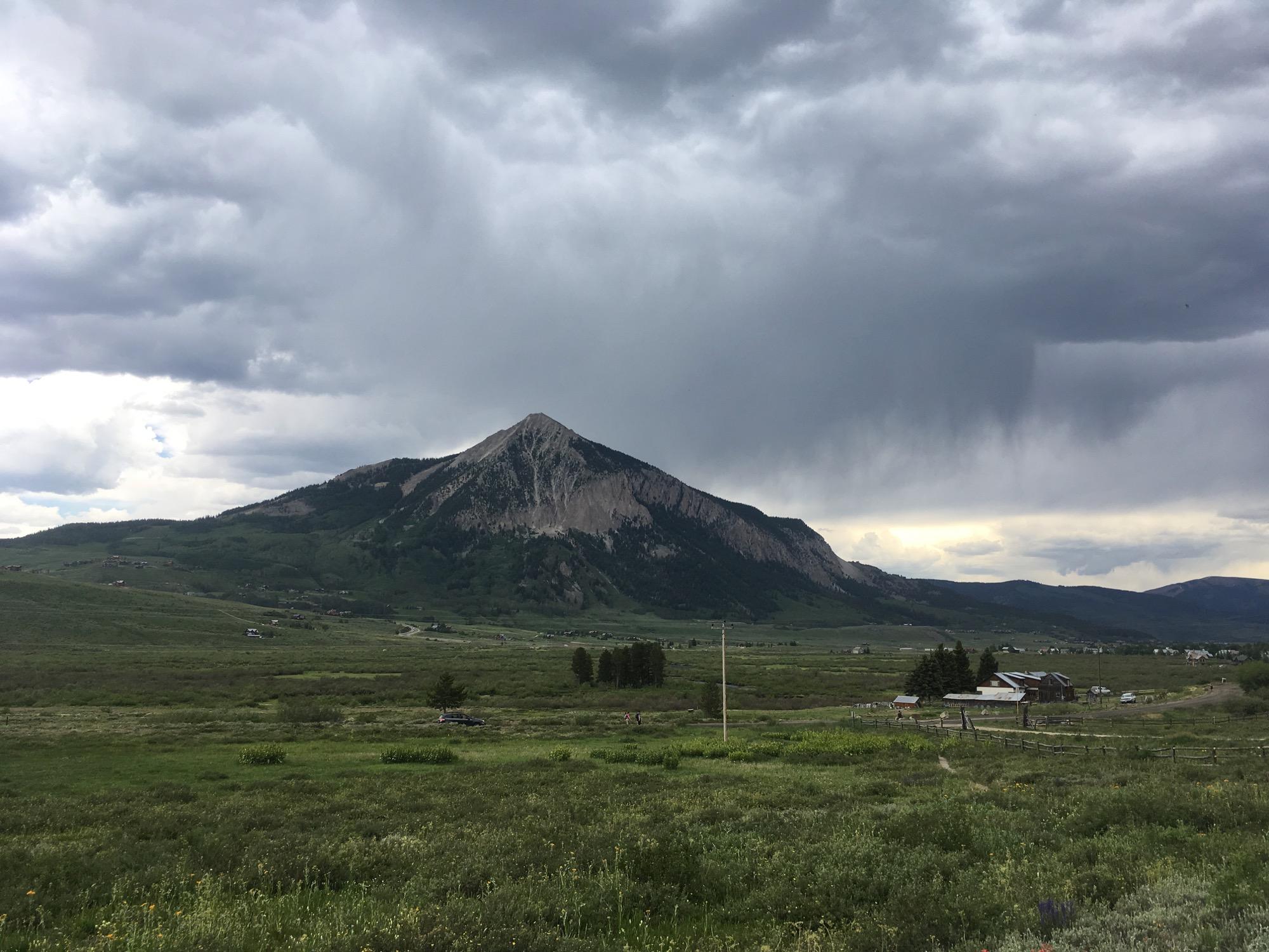 A scenic view of a prominent mountain peak surrounded by green fields under a cloudy sky, with hints of rain in the distance. The landscape features a winding road and a few scattered buildings, adding to the rural charm. Lower Loop mountain bike trail.