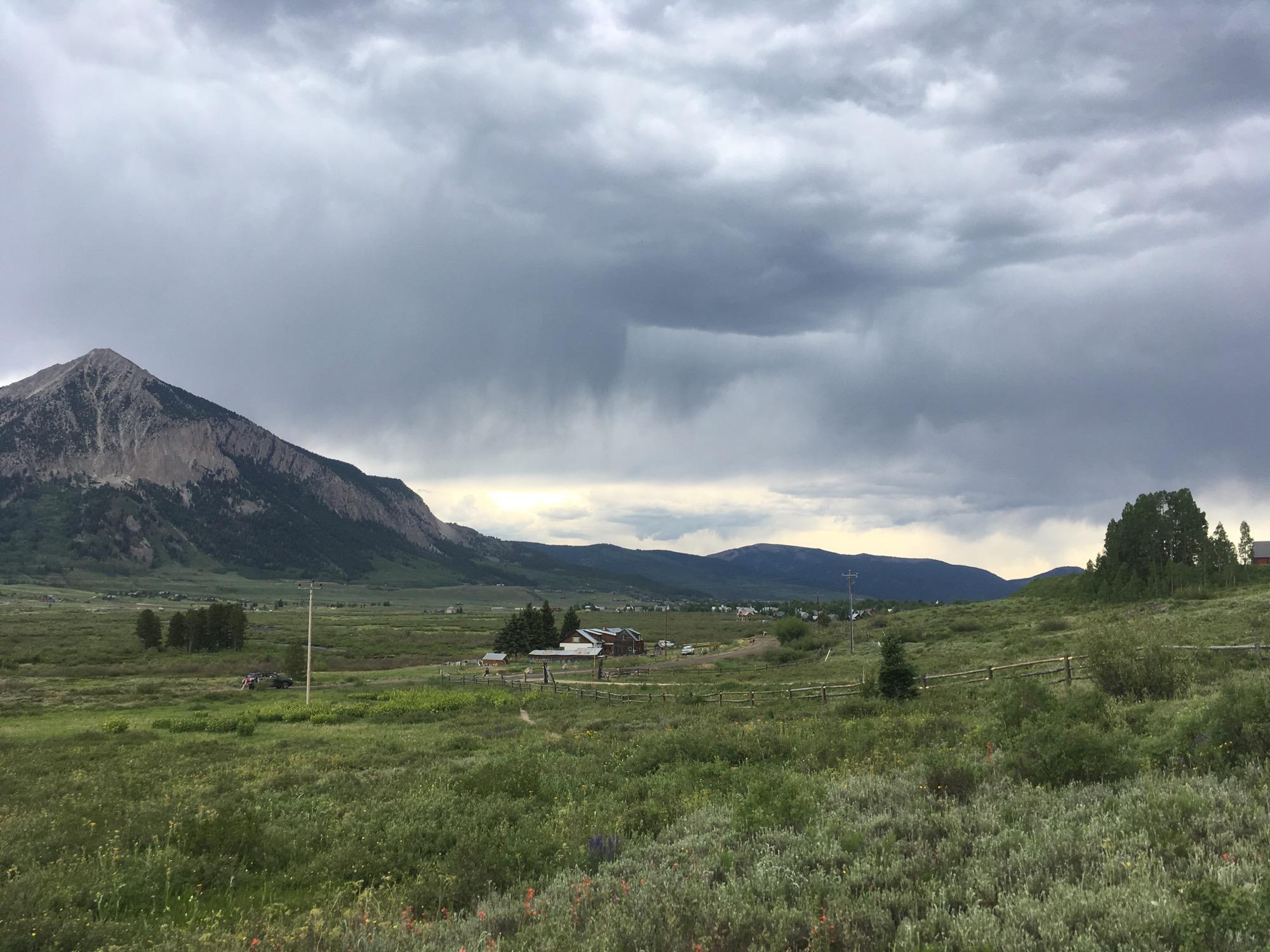 A scenic view of a mountainous landscape under a cloudy sky. A prominent mountain rises in the background, with rugged slopes and patches of greenery. In the foreground, lush fields are dotted with wildflowers and a few structures, hinting at a rural setting. The sky is overcast, suggesting possible rain in the distance. Lower Loop mountain bike trail.