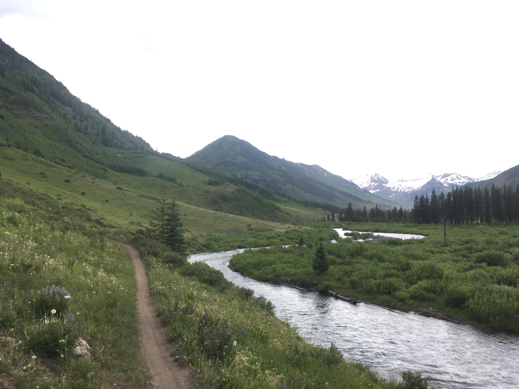 A scenic landscape featuring a winding dirt path alongside a flowing river, surrounded by lush green hills and mountains. Snow-capped peaks are visible in the distance under a cloudy sky, adding to the natural beauty of the scene. Wildflowers line the path, enhancing the vibrant greenery of the area. Lower Loop mountain bike trail.