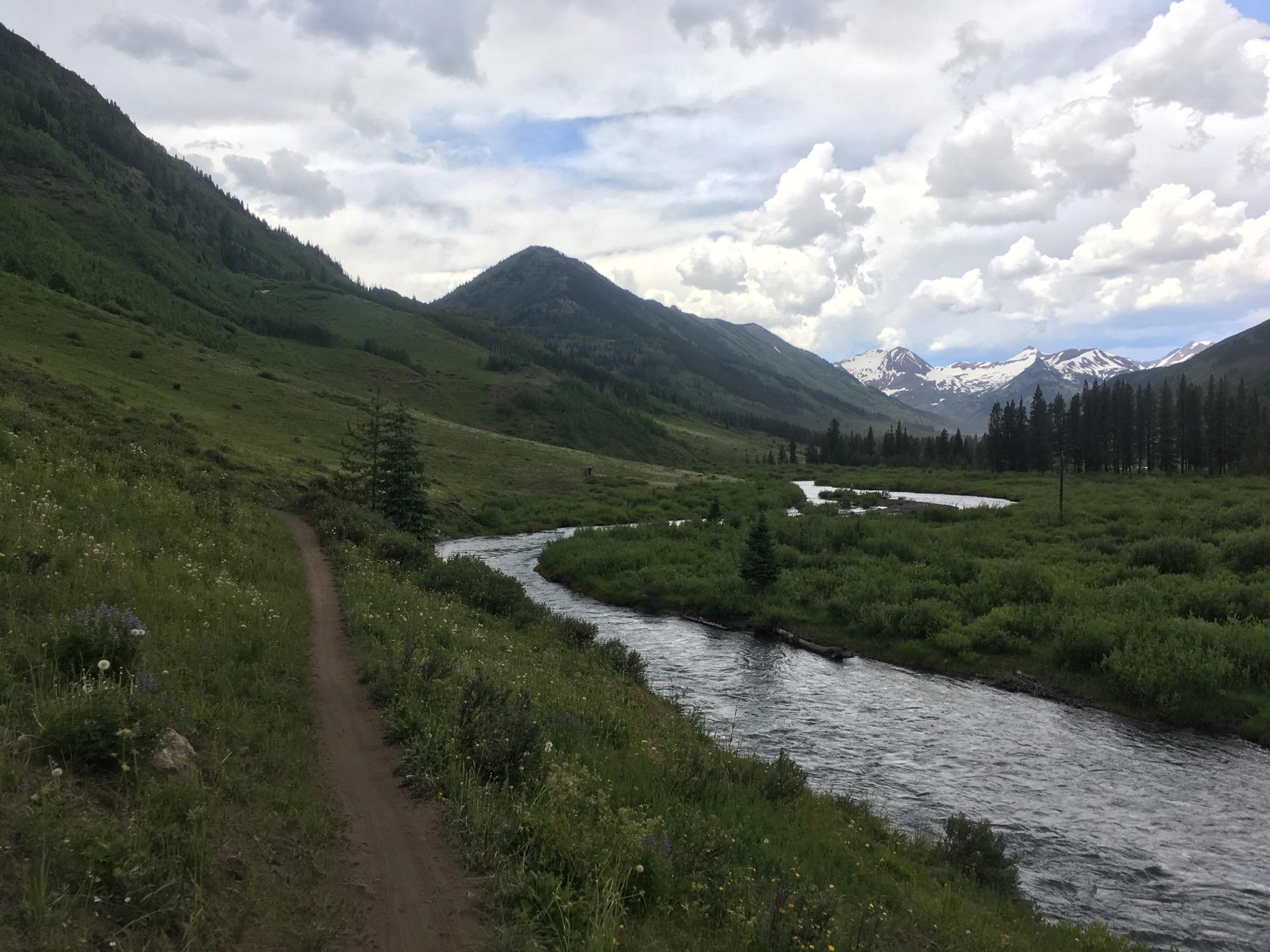 A scenic view of a winding river flowing through a lush green valley, flanked by mountains. The foreground features a dirt path lined with wildflowers, while the background showcases snow-capped peaks under a partly cloudy sky. Lower Loop mountain bike trail.