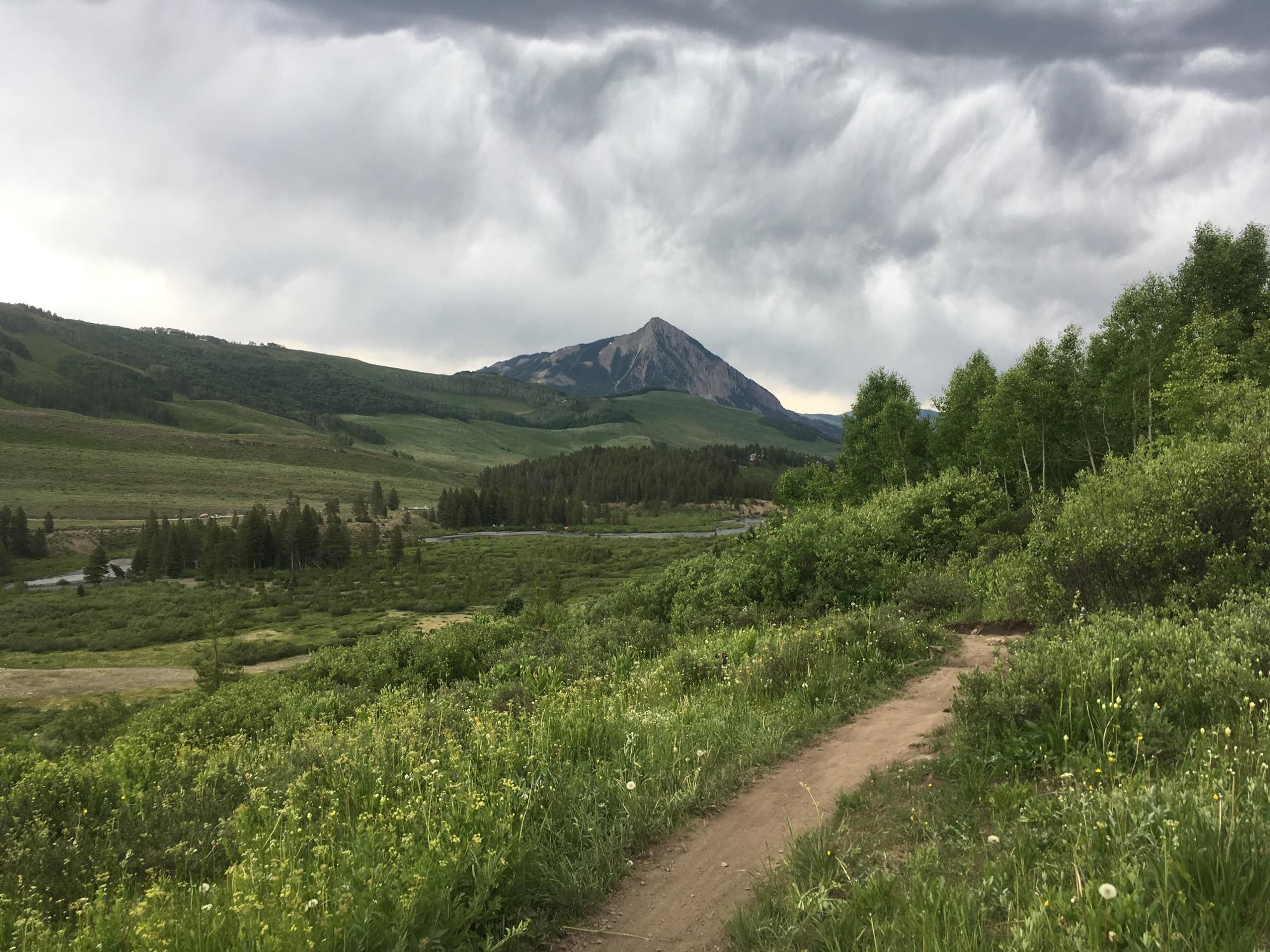 A scenic view of a mountainous landscape under a cloudy sky, featuring lush green valleys and a winding dirt path in the foreground. The prominent peak in the background is partially shrouded in clouds, surrounded by rolling hills and dense clusters of trees. Lower Loop mountain bike trail.