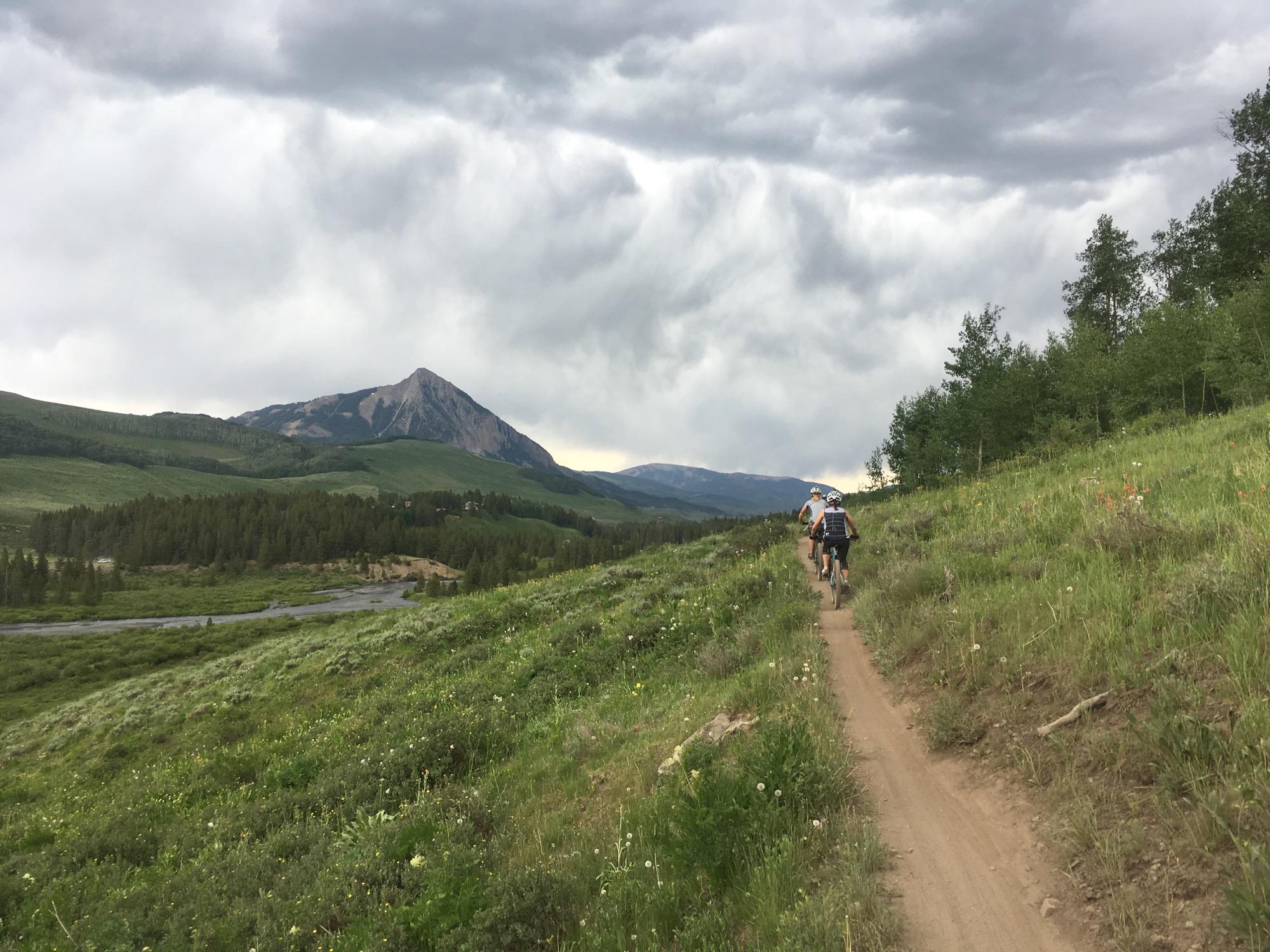 Mountain bikers riding along a dirt trail through a lush, green landscape, with rolling hills and a prominent mountain peak in the background under a cloudy sky. Lower Loop mountain bike trail.