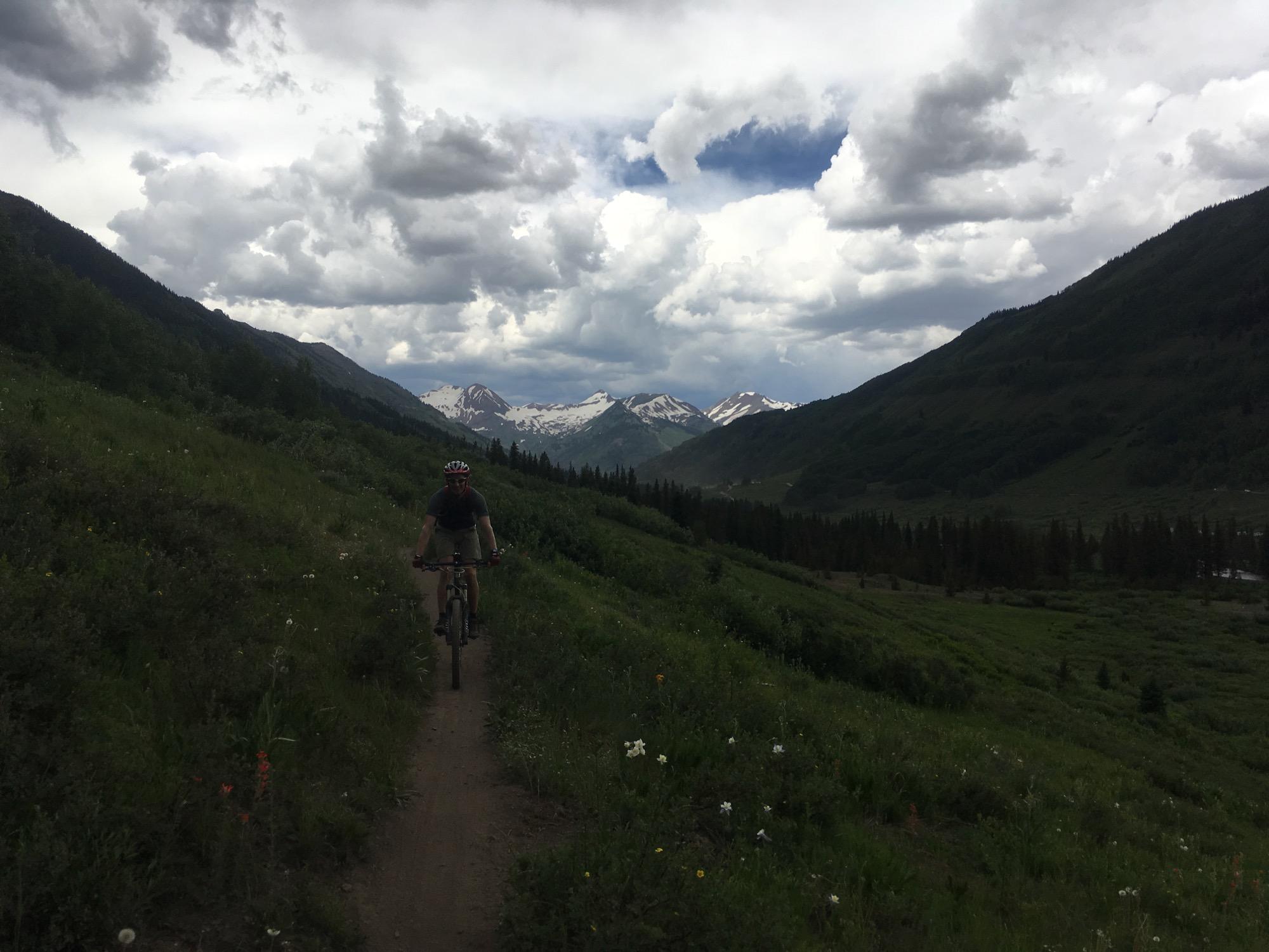 A cyclist rides along a dirt trail in a lush, green valley surrounded by mountains. Dark clouds loom overhead, and snow-capped peaks are visible in the distance, creating a dramatic backdrop. The scene conveys a sense of adventure and natural beauty. Upper Lower Loop mountain bike trail.
