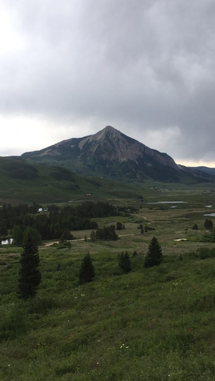 A scenic view of a mountain rising above a lush green valley, with patches of trees and a cloudy sky. The peak of the mountain is prominent and rocky, framed by the soft hills and open meadows below. Lower Loop mountain bike trail.