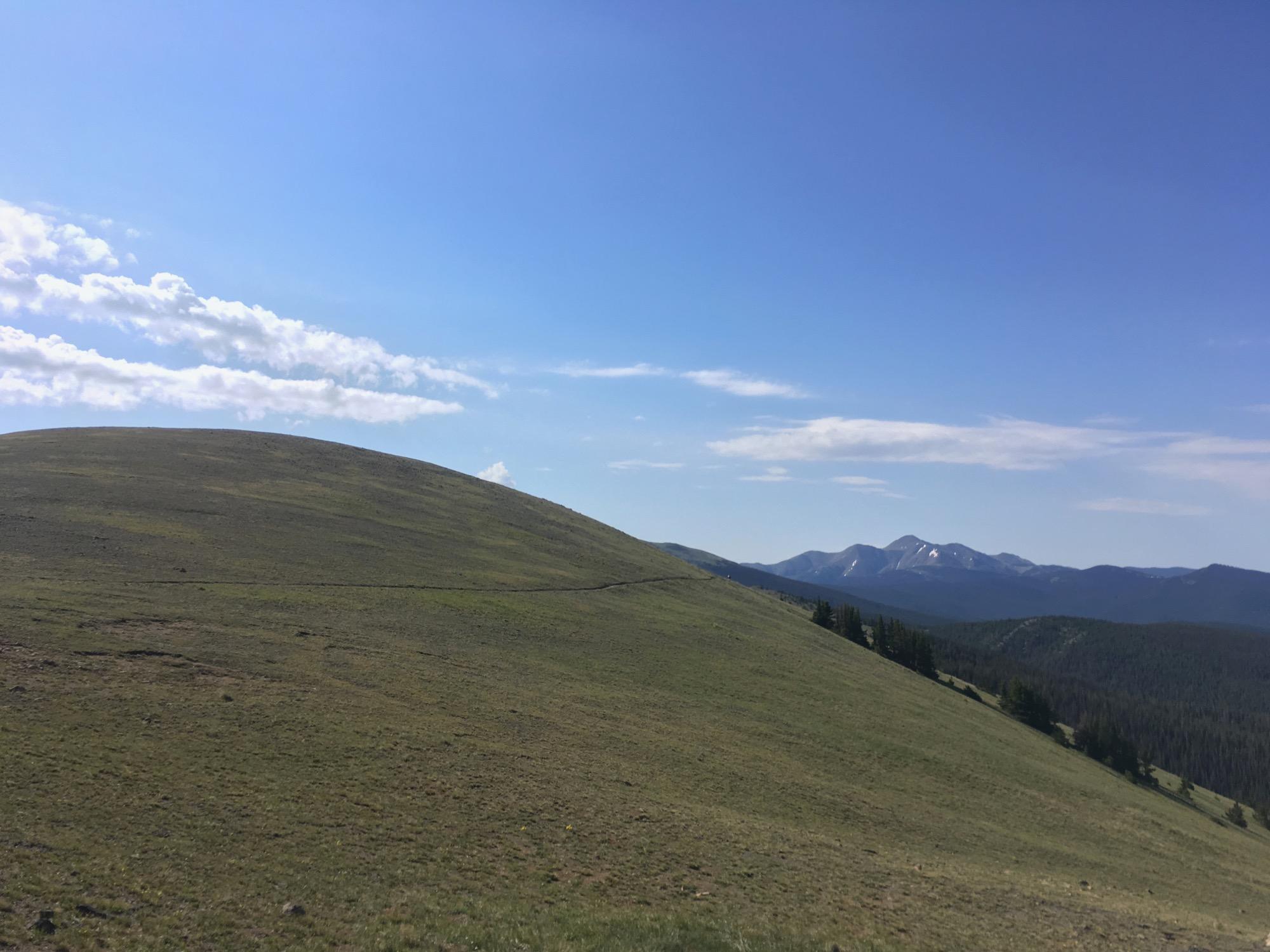 A panoramic view of rolling hills under a clear blue sky, with scattered clouds. In the background, distant mountains are partially covered in snow, and the foreground features a grassy slope. The landscape suggests a serene, natural setting. Monarch Crest Trail mountain bike trail.