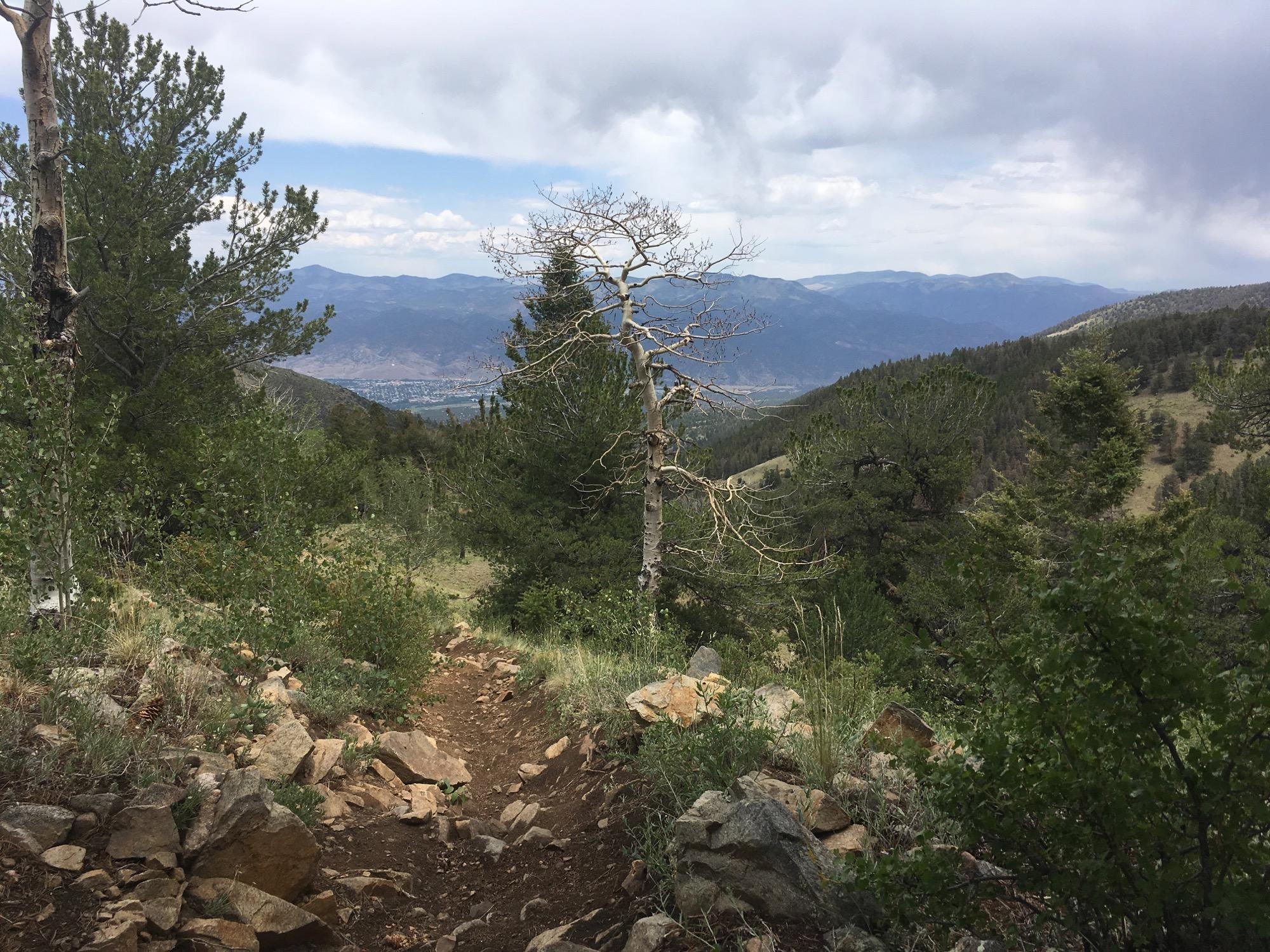 A scenic view of a hiking trail winding through a mountainous landscape, surrounded by lush greenery and rocky terrain. In the distance, rolling hills and mountains are visible under a partly cloudy sky. Rainbow Trail: Hwy 285 to Methodist Mountain Thd mountain bike trail.