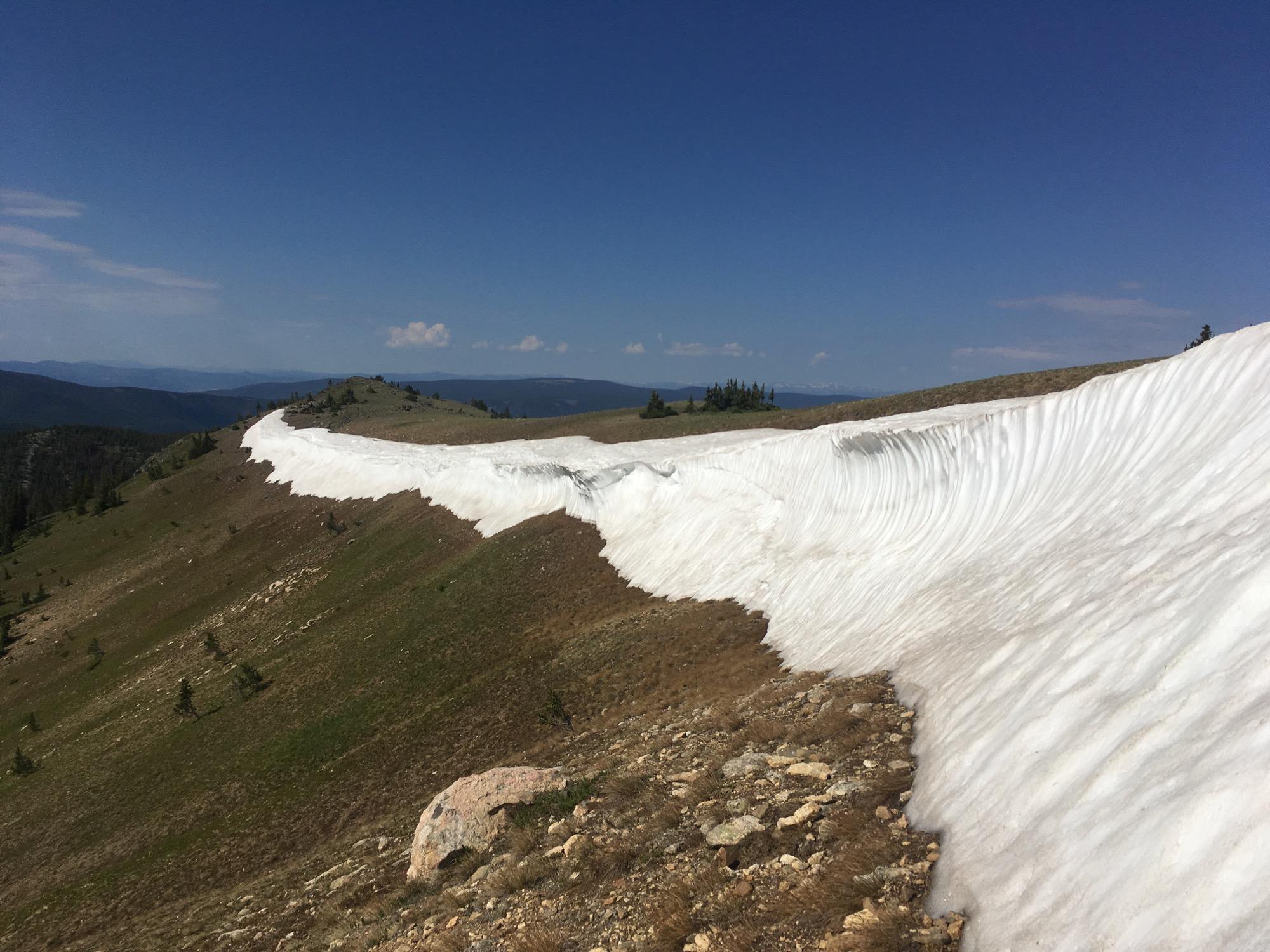 A scenic view of a snowfield on a mountainous landscape, with a clear blue sky above. The snow contrasts with green grassy slopes and rocky terrain, creating a natural boundary along the hilltop. The distant mountains are visible in the background. Monarch Crest Trail mountain bike trail.
