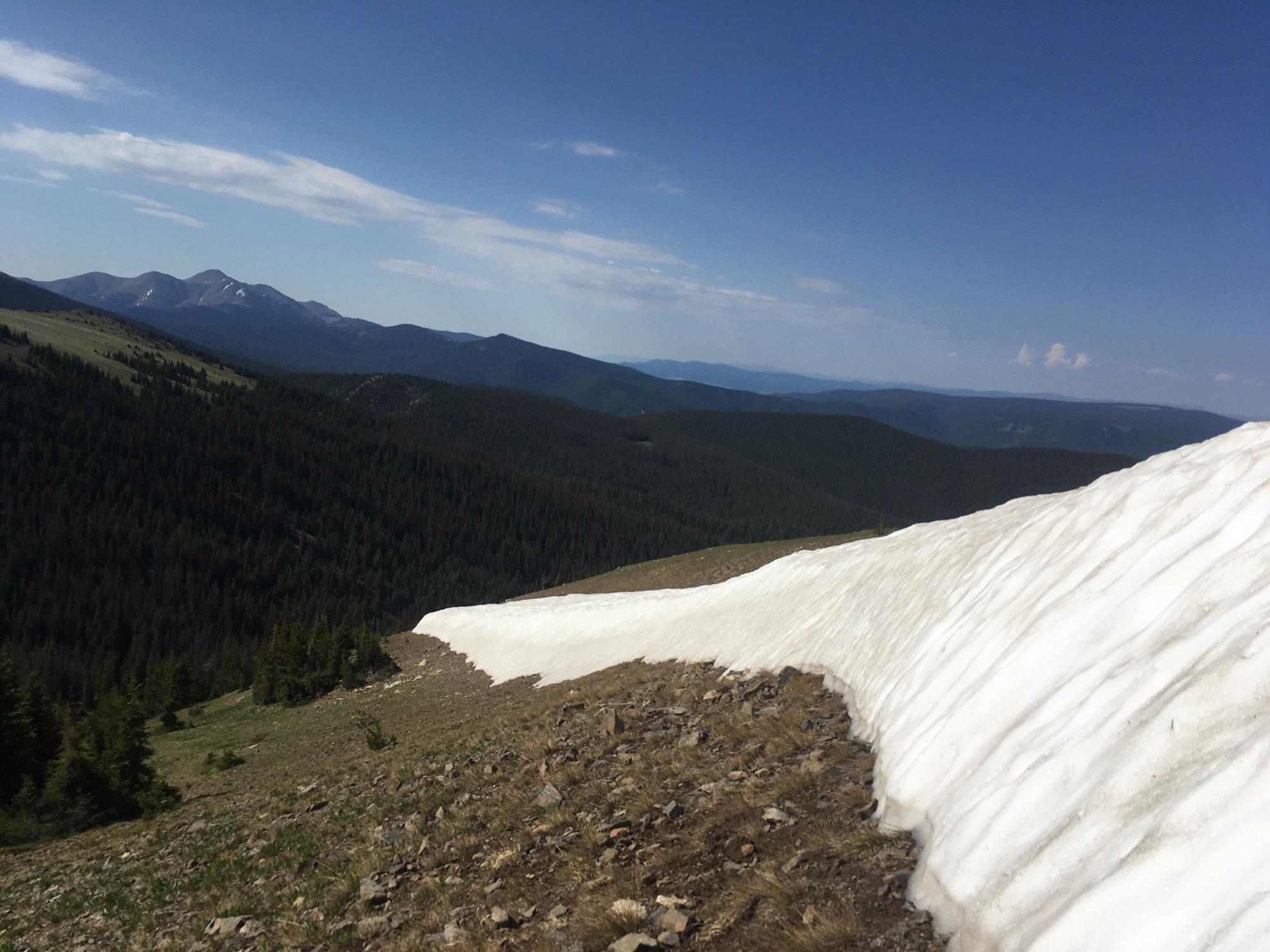 A scenic view of a mountain landscape featuring a snowpack along a ridge, with lush green forests and rolling hills in the background. The sky is clear with a few clouds, and distant mountains are visible on the horizon. Monarch Crest Trail mountain bike trail.