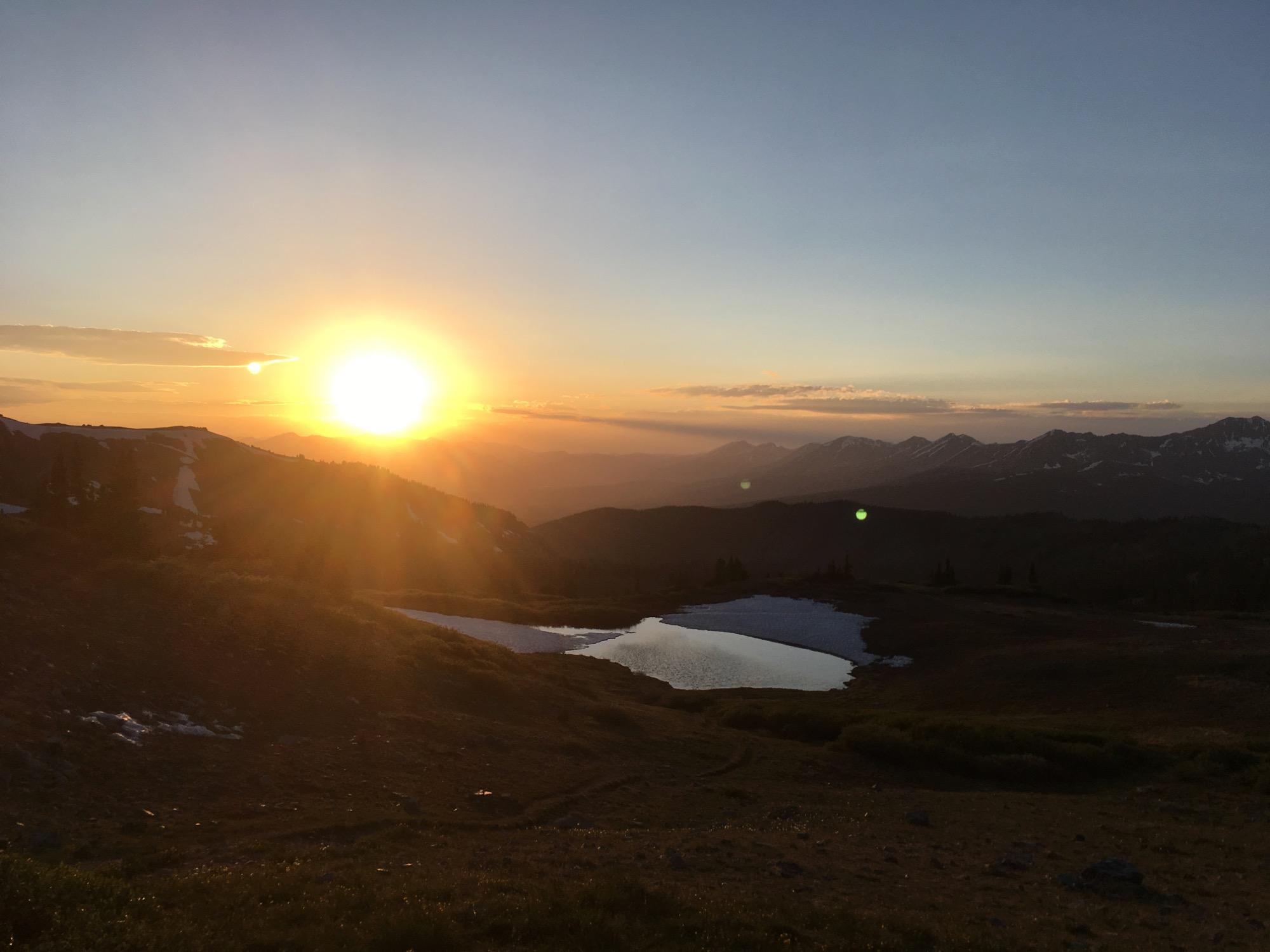 A breathtaking sunset over mountainous terrain, with the sun casting a warm glow across the landscape. In the foreground, a small, calm pond reflects the colors of the sky, while patches of snow are visible on the mountains. The scene showcases a serene and picturesque natural setting at dusk. Cottonwood Pass Road mountain bike trail.