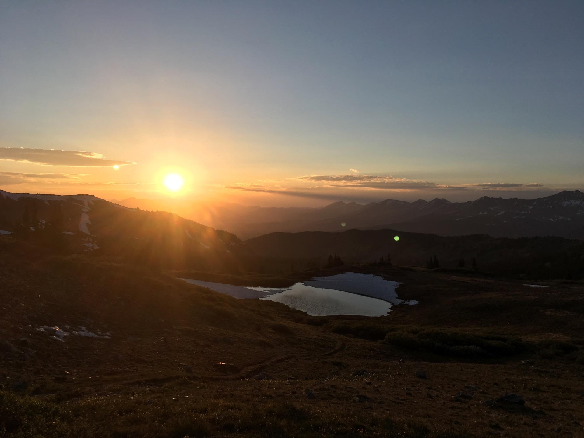 Sunset over a mountainous landscape, with the sun casting a warm golden glow on the horizon. In the foreground, a serene pond reflects the colors of the sky, while rugged mountain peaks and patches of snow are visible in the background. The scene conveys a tranquil and picturesque view of nature. Cottonwood Pass Road mountain bike trail.