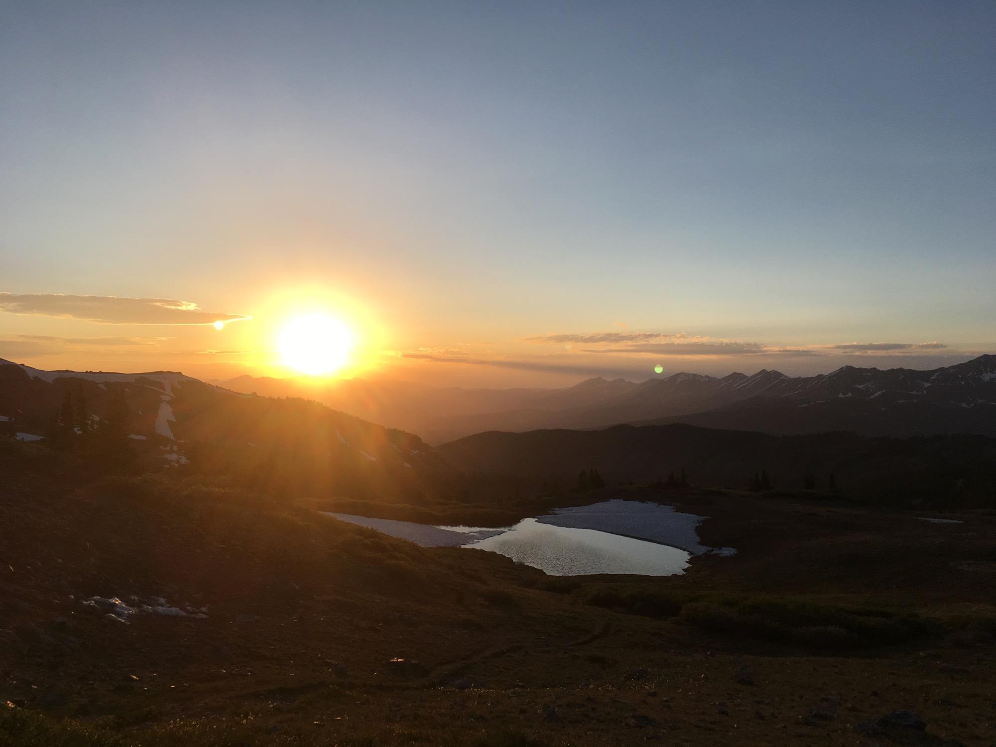Alt text: A breathtaking sunset over a mountainous landscape, with the sun casting a golden glow on the peaks and a small pond reflecting the light. Snow patches are visible on the mountains, and a clear sky enhances the serene atmosphere. Cottonwood Pass Road mountain bike trail.
