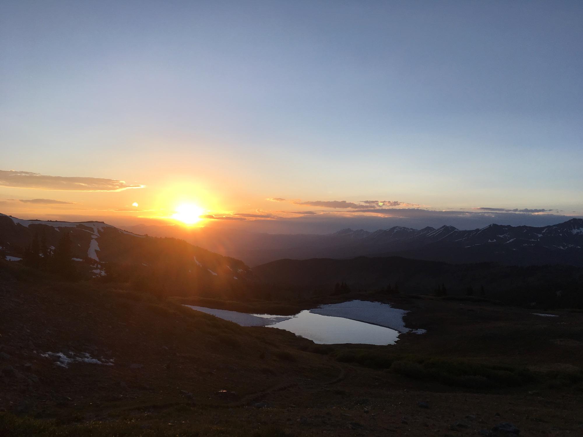 Sunset over a mountain landscape, featuring a bright orange sun dipping below the horizon, casting warm light across snow-capped peaks and a small serene lake reflecting the colors of the sky. Cottonwood Pass Road mountain bike trail.