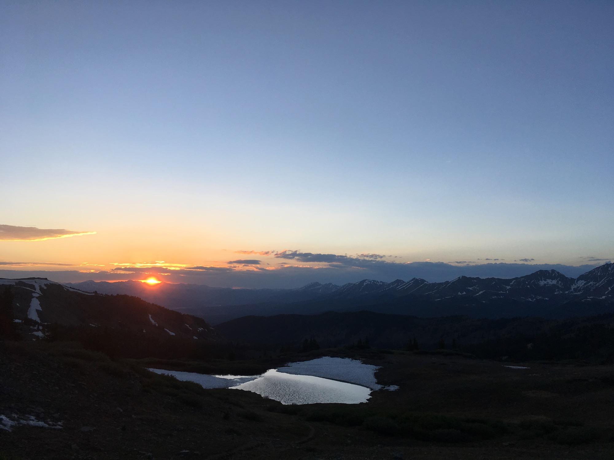 Sunset over a mountain landscape, with the sun partially hidden behind distant peaks. A calm lake reflects the colorful hues of the sky, which transitions from deep blue to warm orange and yellow. Snow can be seen on the mountains in the background, and the scene conveys a sense of tranquility and natural beauty. Cottonwood Pass Road mountain bike trail.
