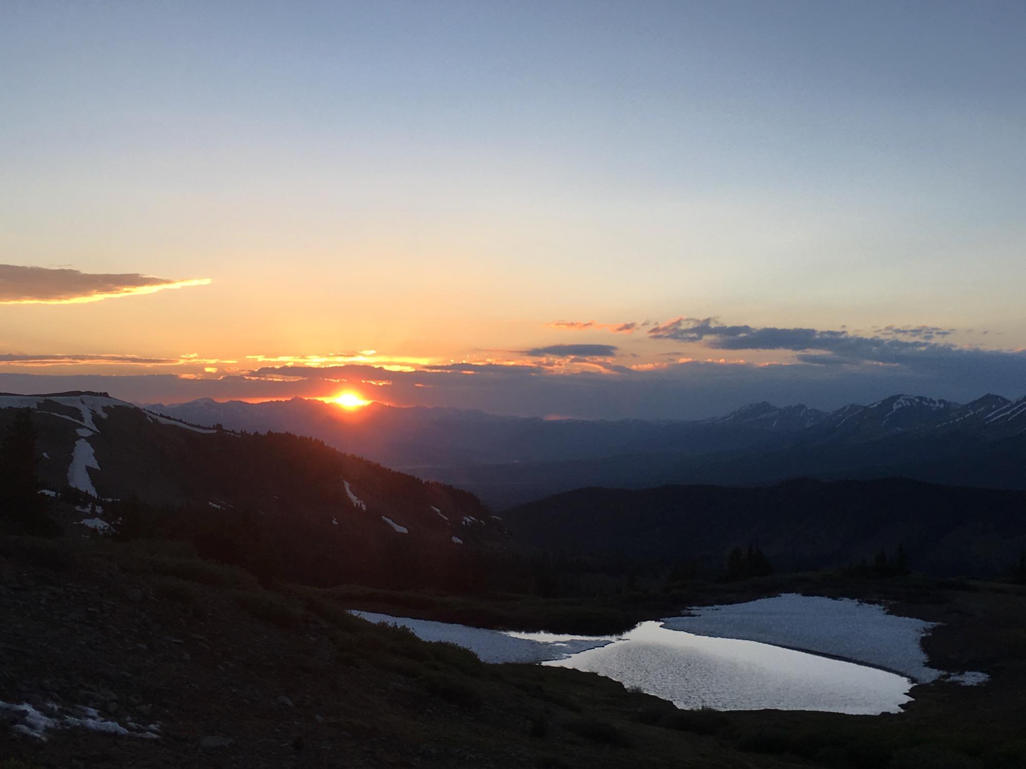 A scenic sunset over a mountainous landscape, with the sun dipping below the horizon and casting a warm glow in the sky. Snow-capped peaks are visible in the distance, while a small body of water reflects the colors of the sky. The foreground features rugged terrain with patches of snow and greenery. Cottonwood Pass Road mountain bike trail.