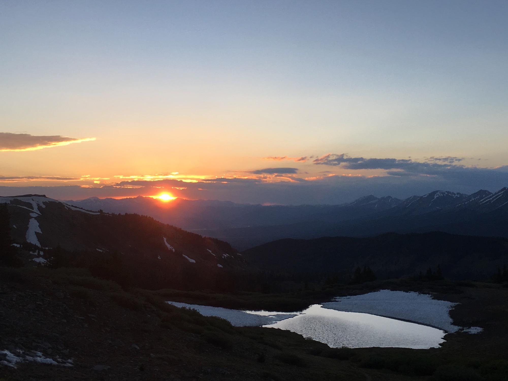 Sunset over a mountainous landscape, with the sun dipping below the horizon and illuminating the sky in shades of orange and pink. In the foreground, a small pond reflects the colors of the sunset, surrounded by lush greenery and patches of snow on the mountainside. The distant peaks are silhouetted against the colorful sky. Cottonwood Pass Road mountain bike trail.