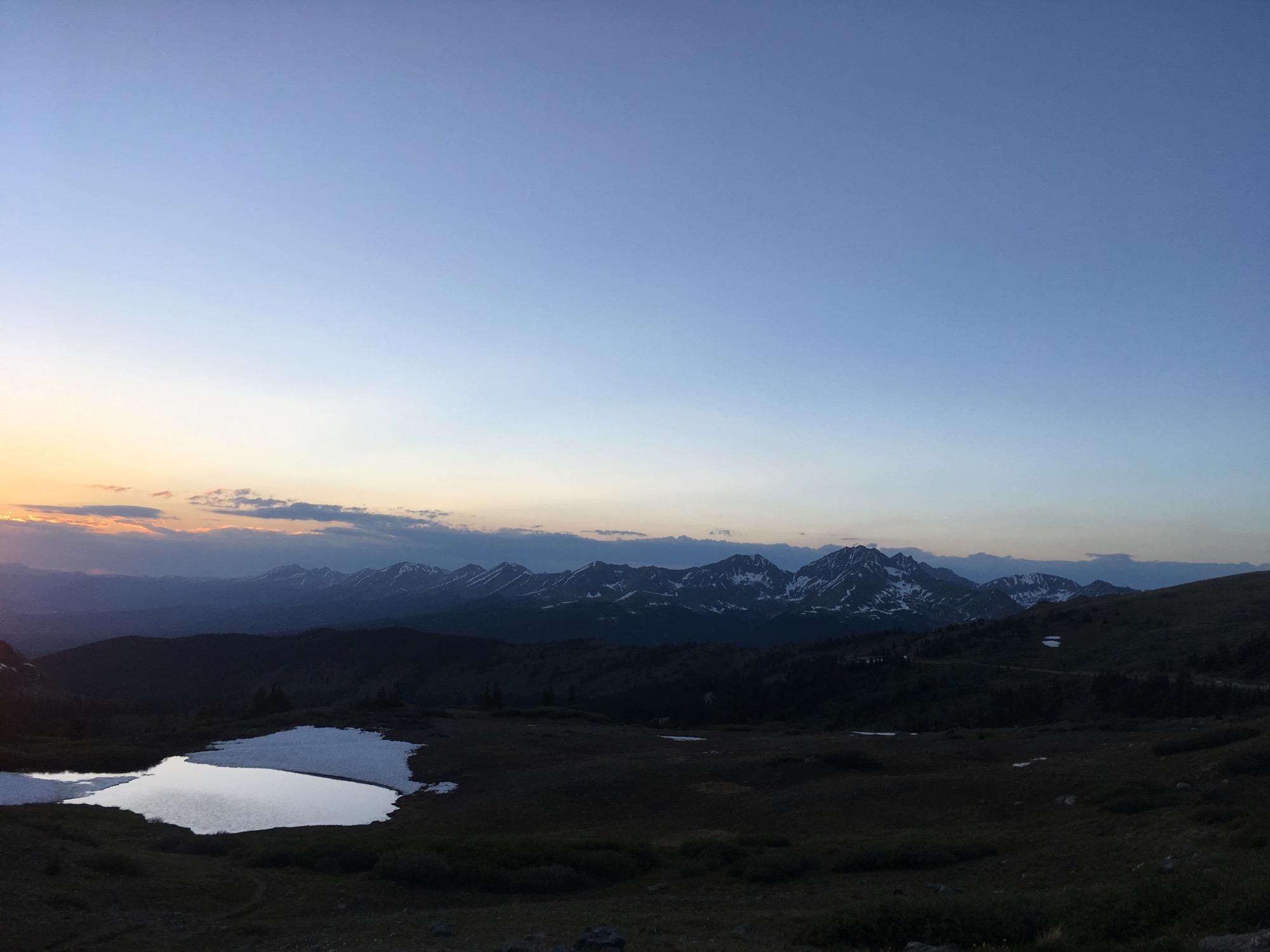 A serene mountain landscape at sunset, featuring a clear sky transitioning from orange to blue. Snow-capped peaks stretch across the horizon, with a small reflective pond in the foreground. Lush green meadows and patches of snow add to the scenic beauty of the high-altitude setting. Cottonwood Pass Road mountain bike trail.